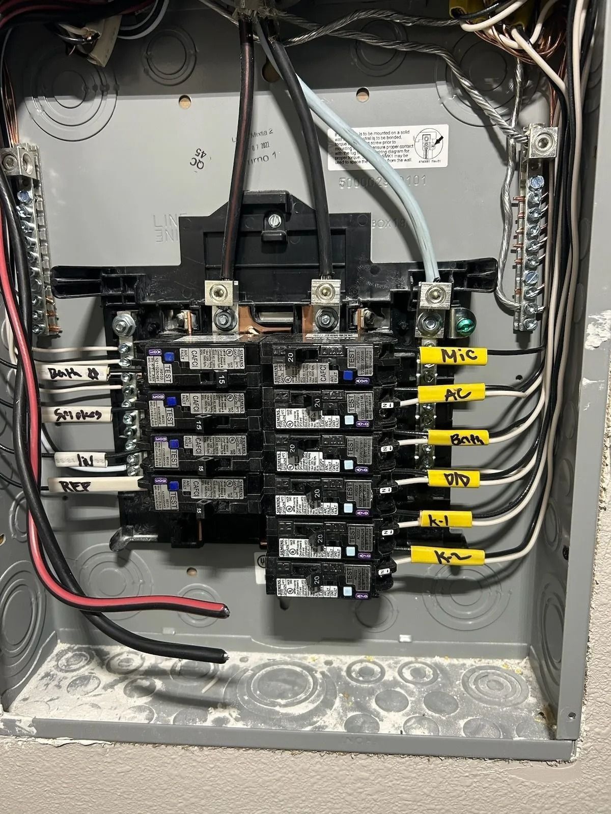 Electrical panel with circuit breakers and wiring inside a gray metal box. Labeled circuits include