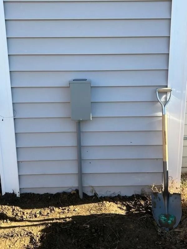 Gray electrical box on a metal pole next to a house with gray siding. A shovel leans against the house.