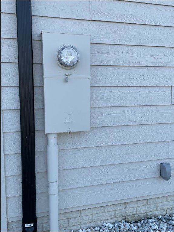 Electrical meter and panel on a light-colored, wood-sided building exterior, with a black downspout.