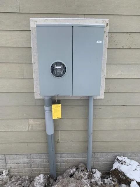 Gray electrical meter box on a beige house exterior with gray conduit pipes and snow.
