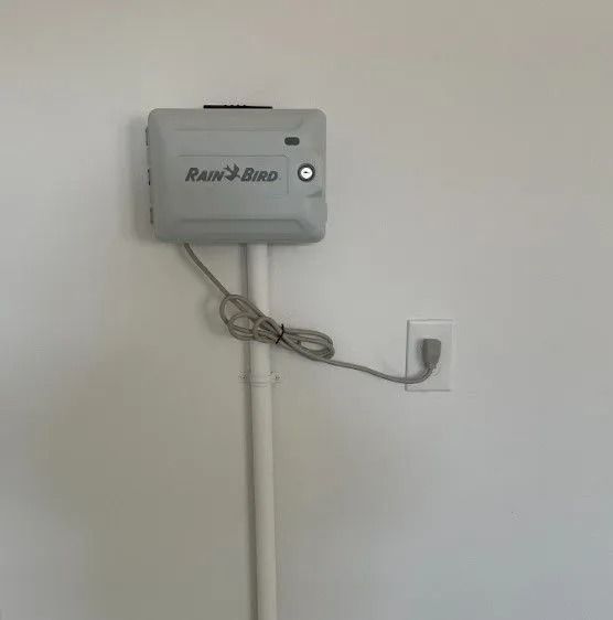 Rain Bird irrigation controller mounted on a white wall, plugged into an outlet.