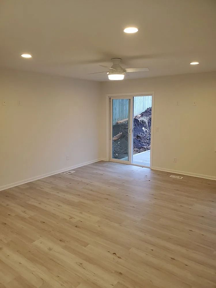 Empty room with light wood floor, white walls, recessed lights, ceiling fan, and sliding glass door.