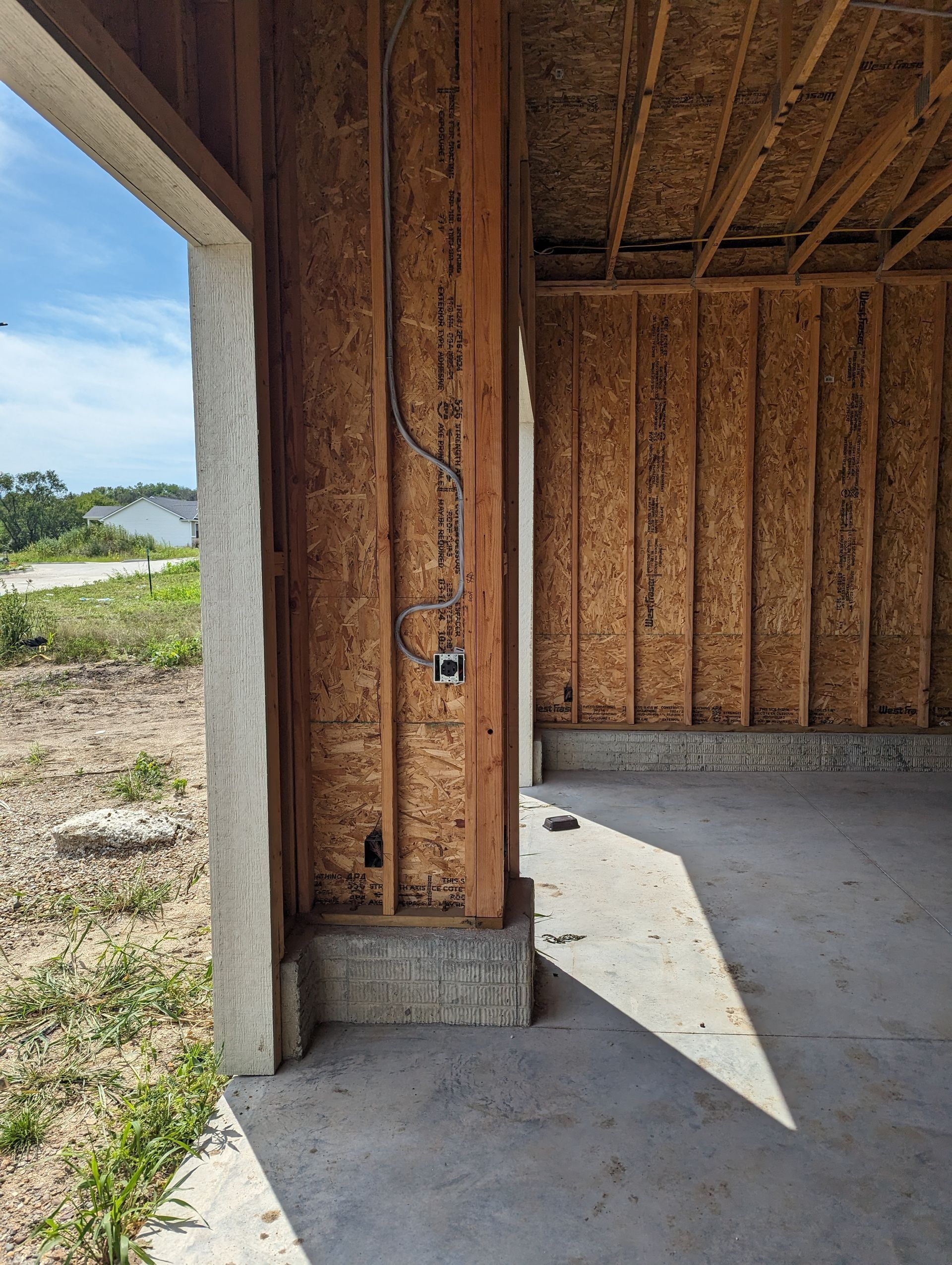 Unfinished building exterior: wooden framing, electrical outlet, concrete floor and supporting column. Blue sky visible.