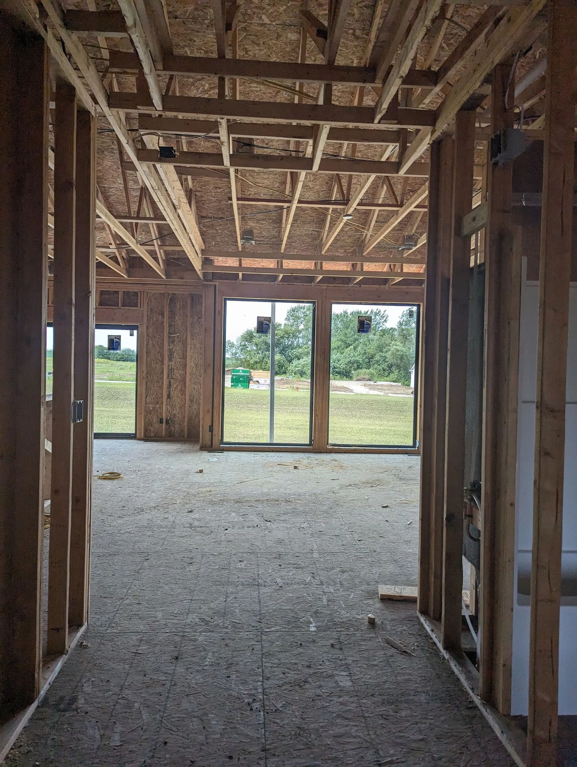 Interior view of a building under construction, framed with wood. Large windows look out to green trees and grass.