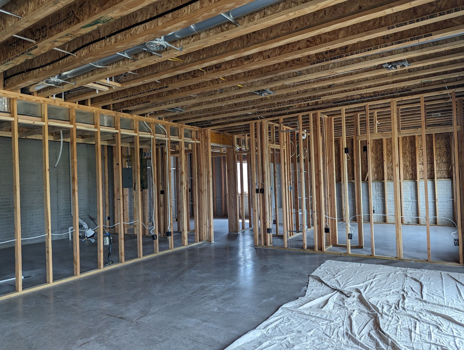 Interior view of a building in the framing stage. Exposed wooden studs and joists, concrete floor, white tarp.