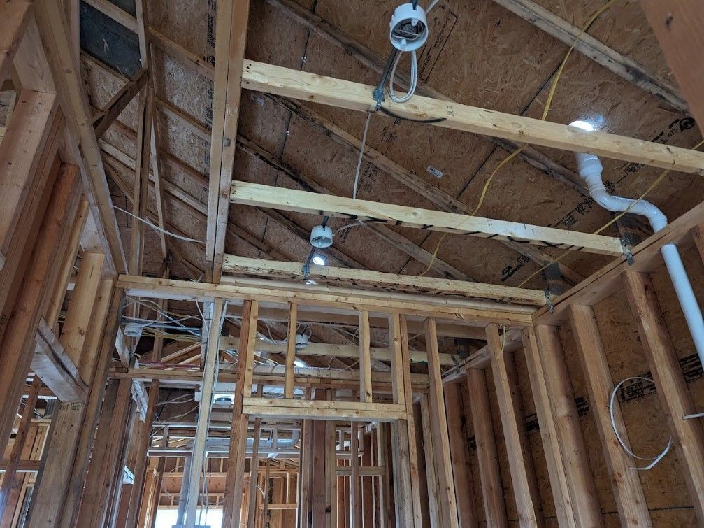 Interior of a house under construction; exposed wooden framing, ceiling lights, and electrical wiring are visible.