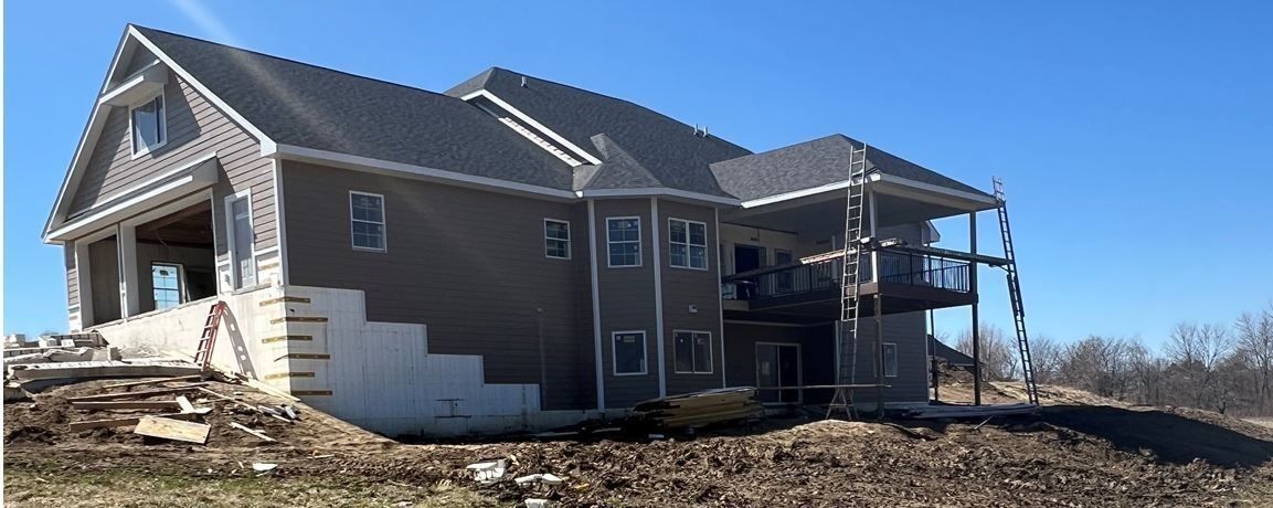 A large, multi-level house under construction with gray siding and dark roof under a clear blue sky.
