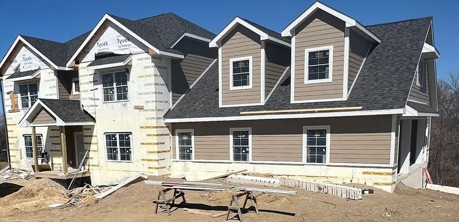House under construction with tan siding and dark gray roof against a clear blue sky.