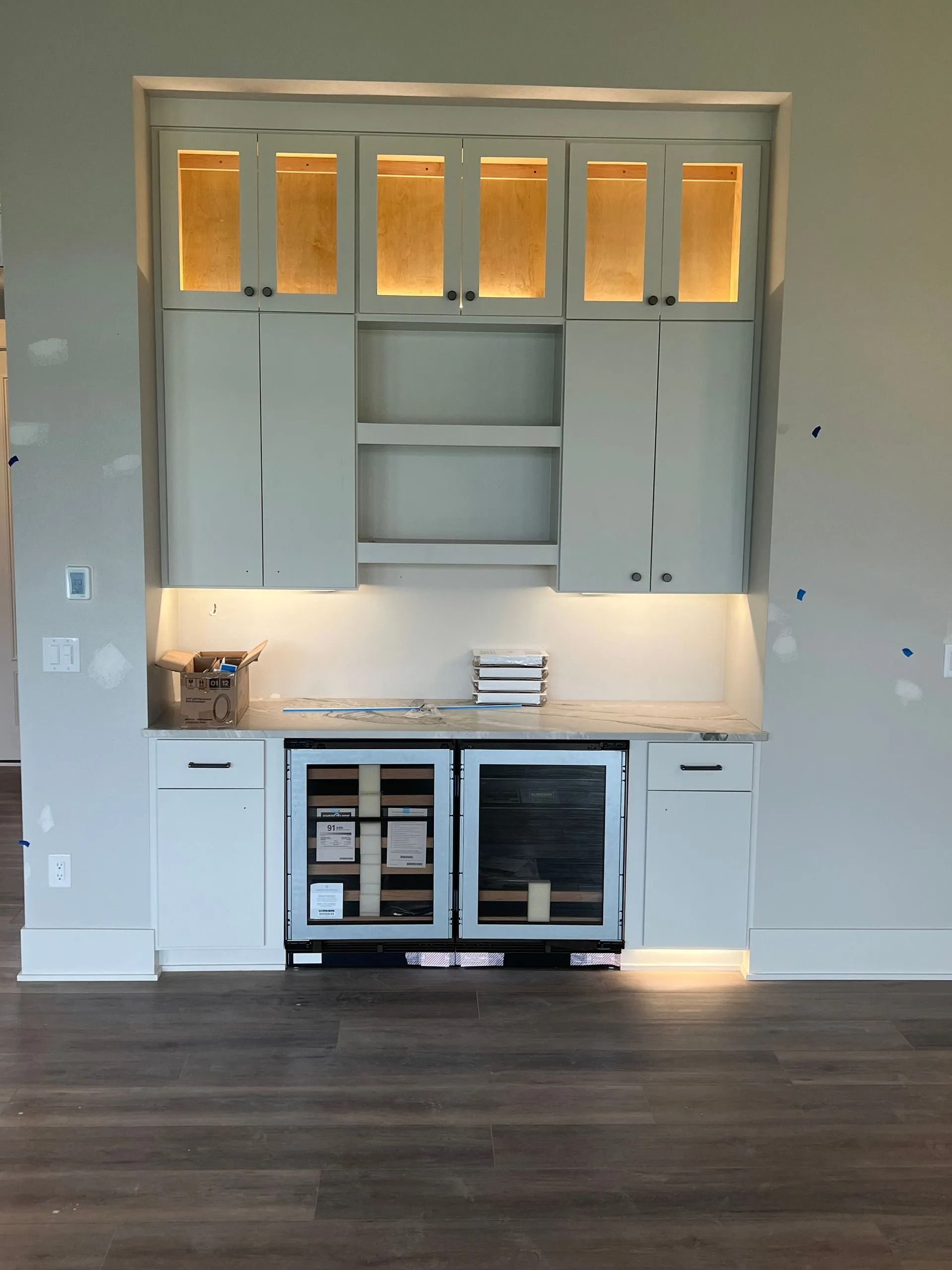 Built-in bar with pale blue cabinets, open shelving, wine fridge, and overhead lighting in a room with wood flooring.