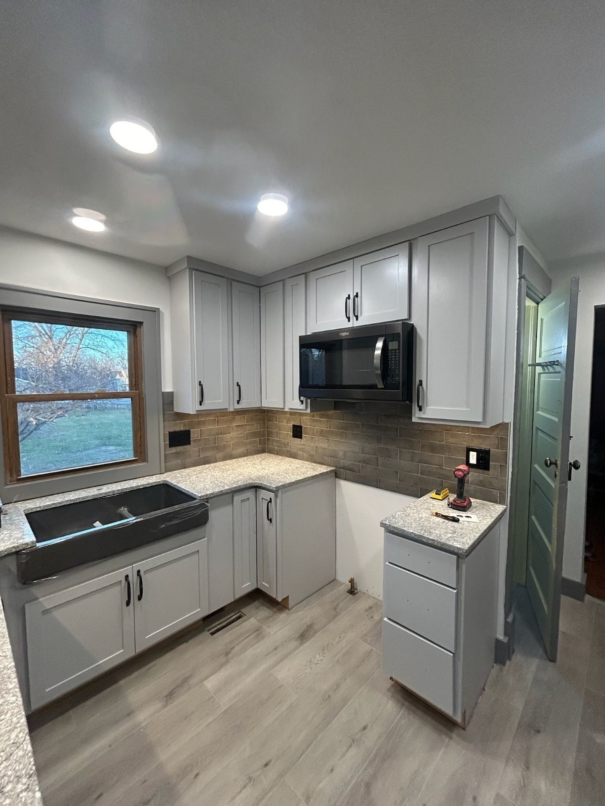 Light gray kitchen with cabinets, backsplash, black microwave, and double sink.