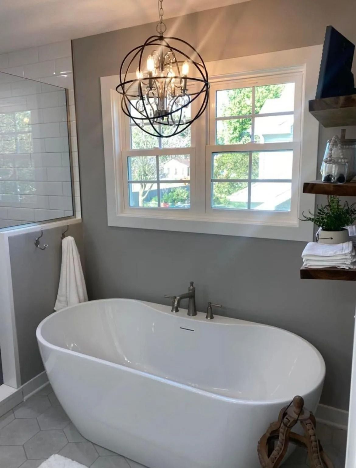 Bathroom with a white freestanding tub, window, and decorative lighting. Gray walls, and wooden shelves are visible.