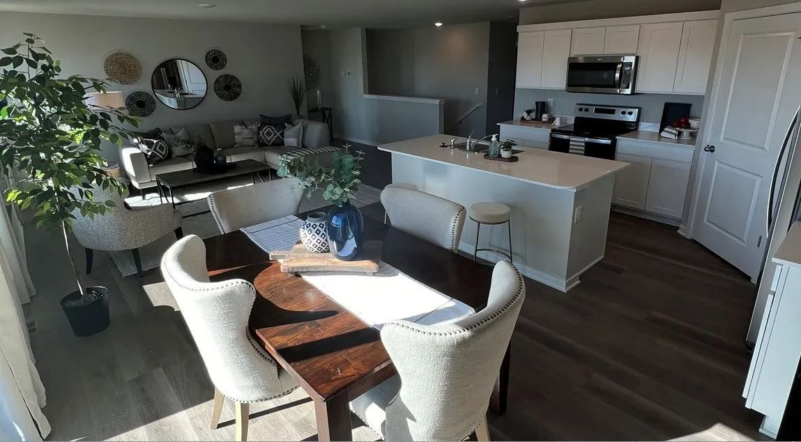 Open-plan living space: kitchen island, dining table, sofa, potted plant, neutral tones, hardwood floors.