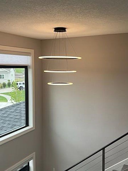 Modern pendant light fixture with three illuminated rings in a neutral-toned hallway, near a window and stairs.