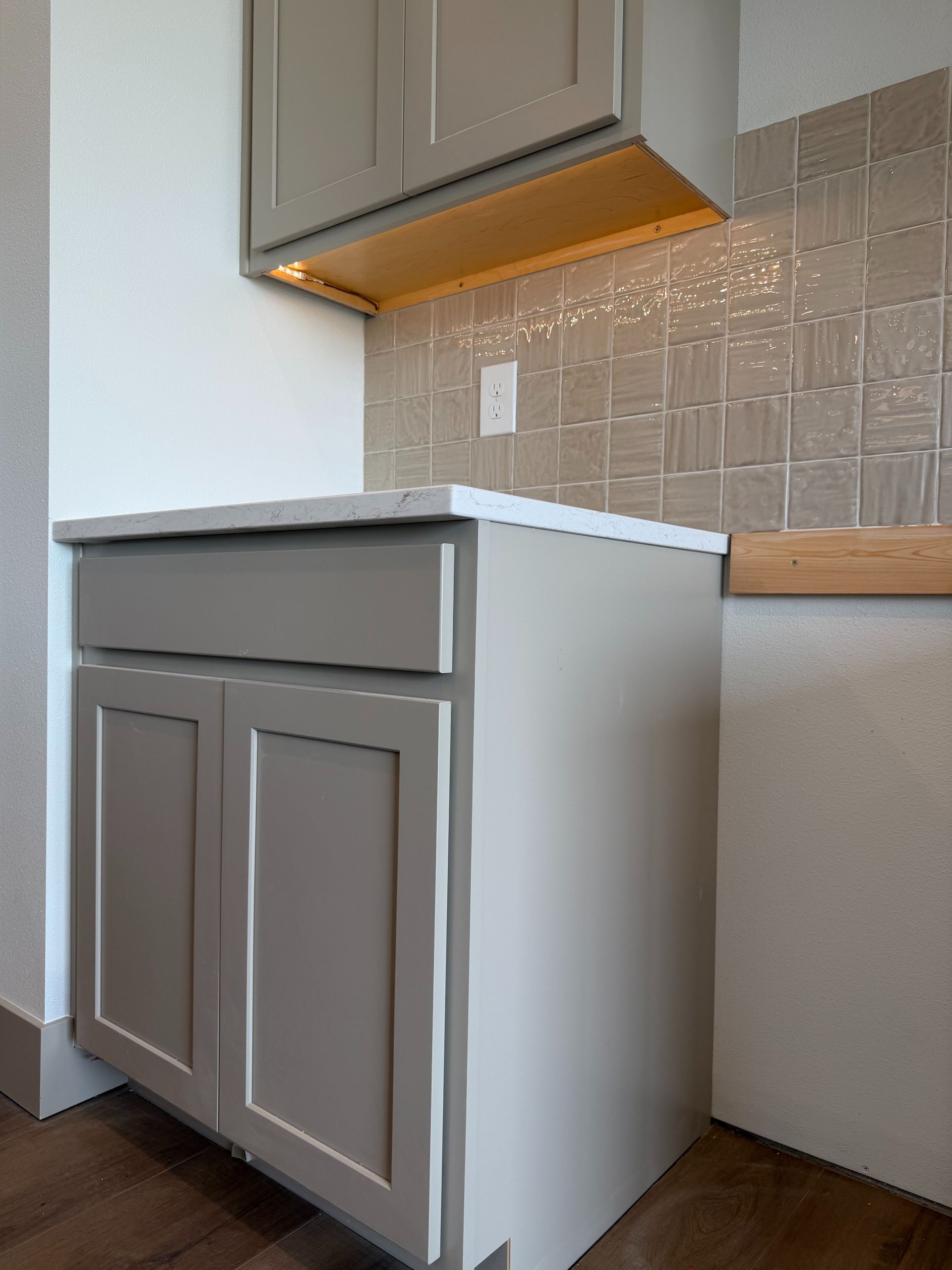 Gray kitchen cabinet with countertop, tan backsplash, and wooden floor in a corner.