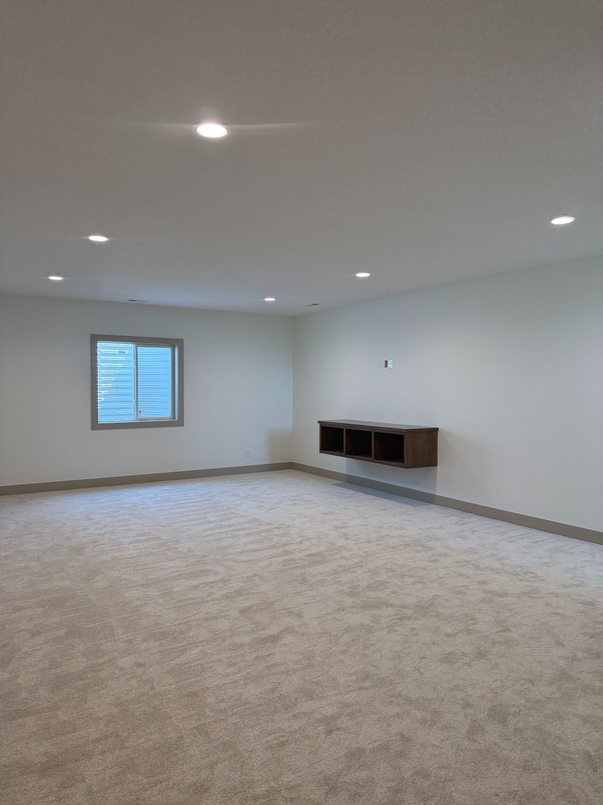 Empty carpeted room with white walls, recessed lights, a small window, and a wall-mounted shelf.