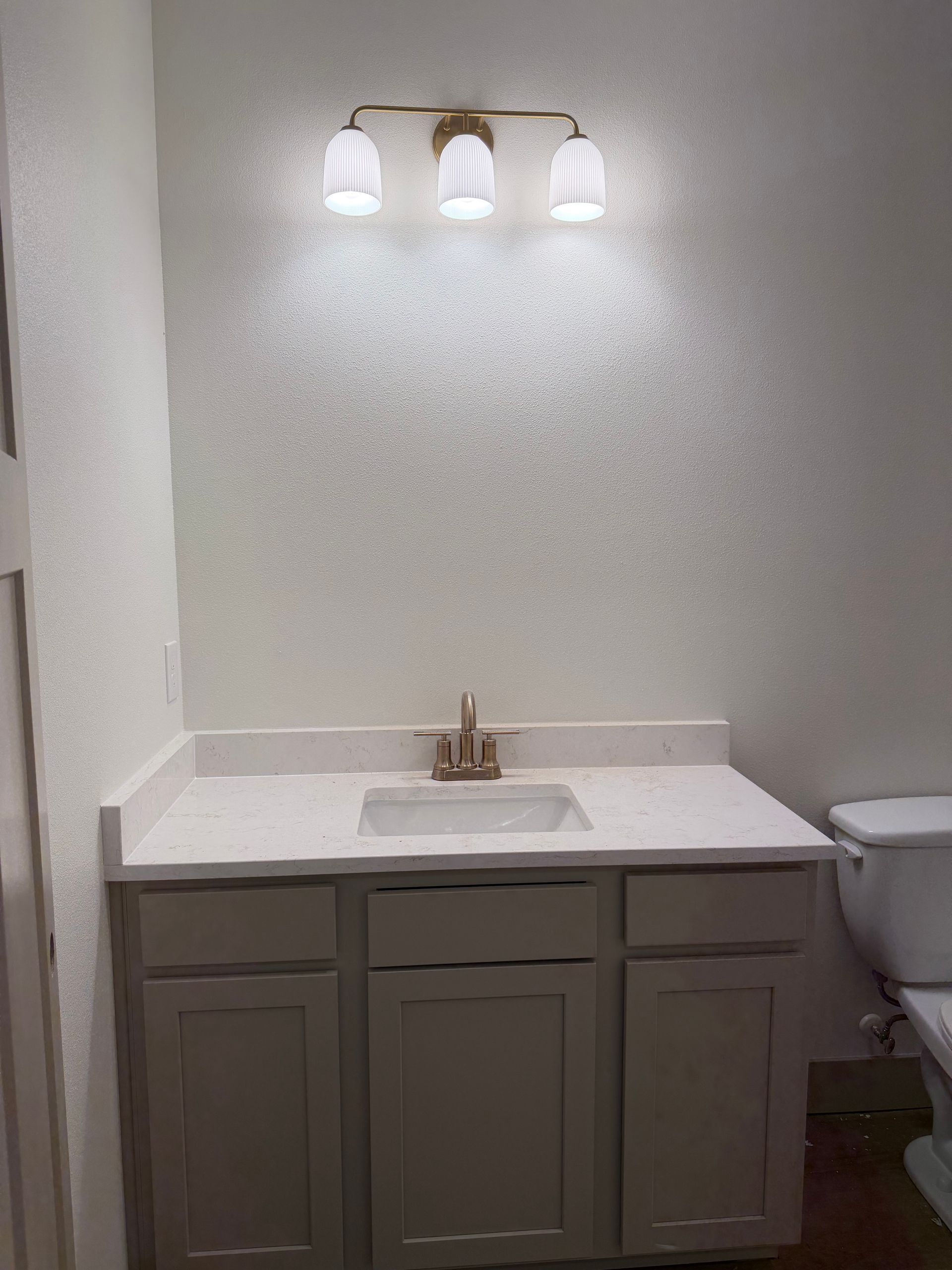 Bathroom vanity with white countertop, gray cabinet, and three-light wall fixture beside a toilet.