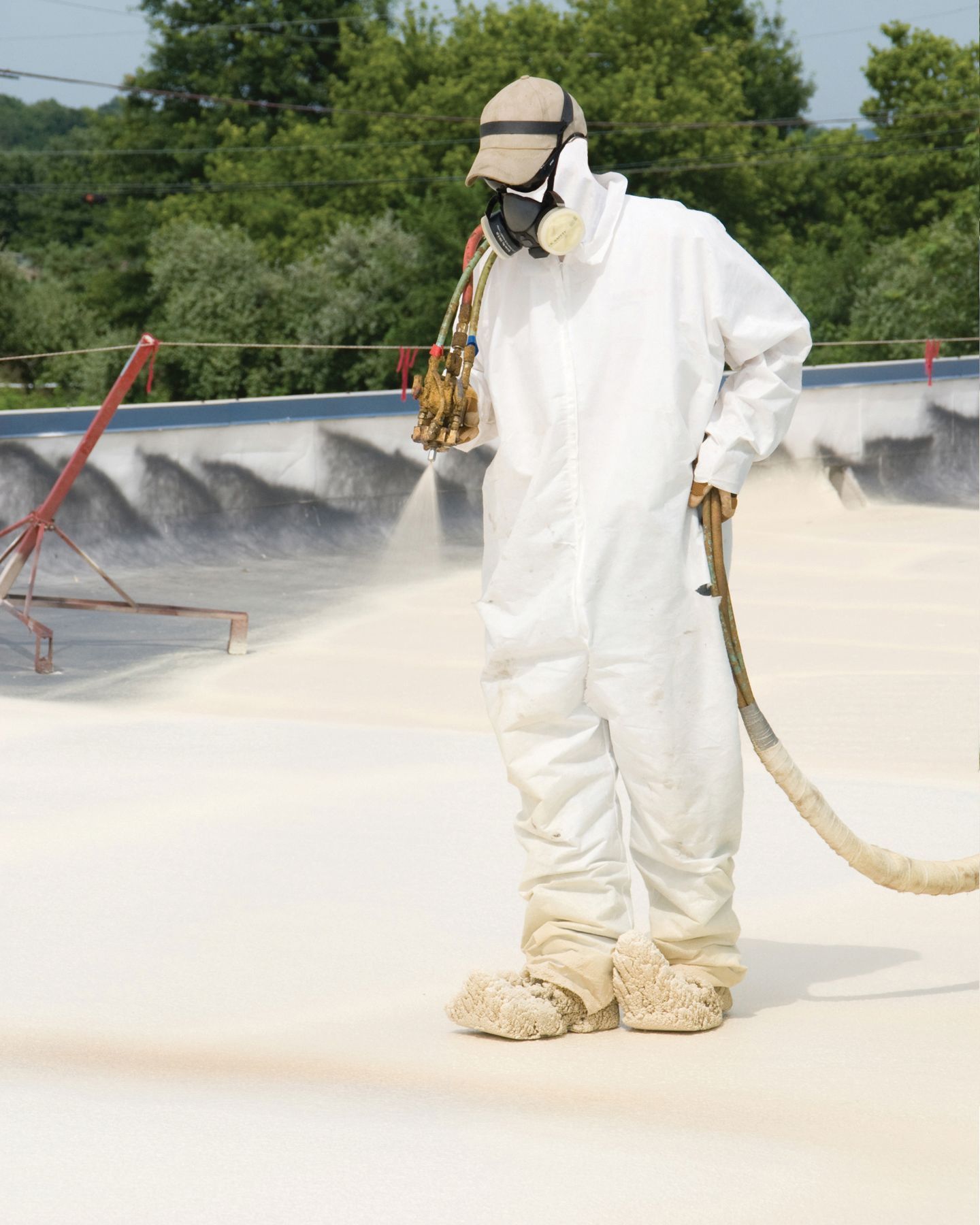 A worker in a white protective suit and mask sprays a coating onto a flat roof outdoors.