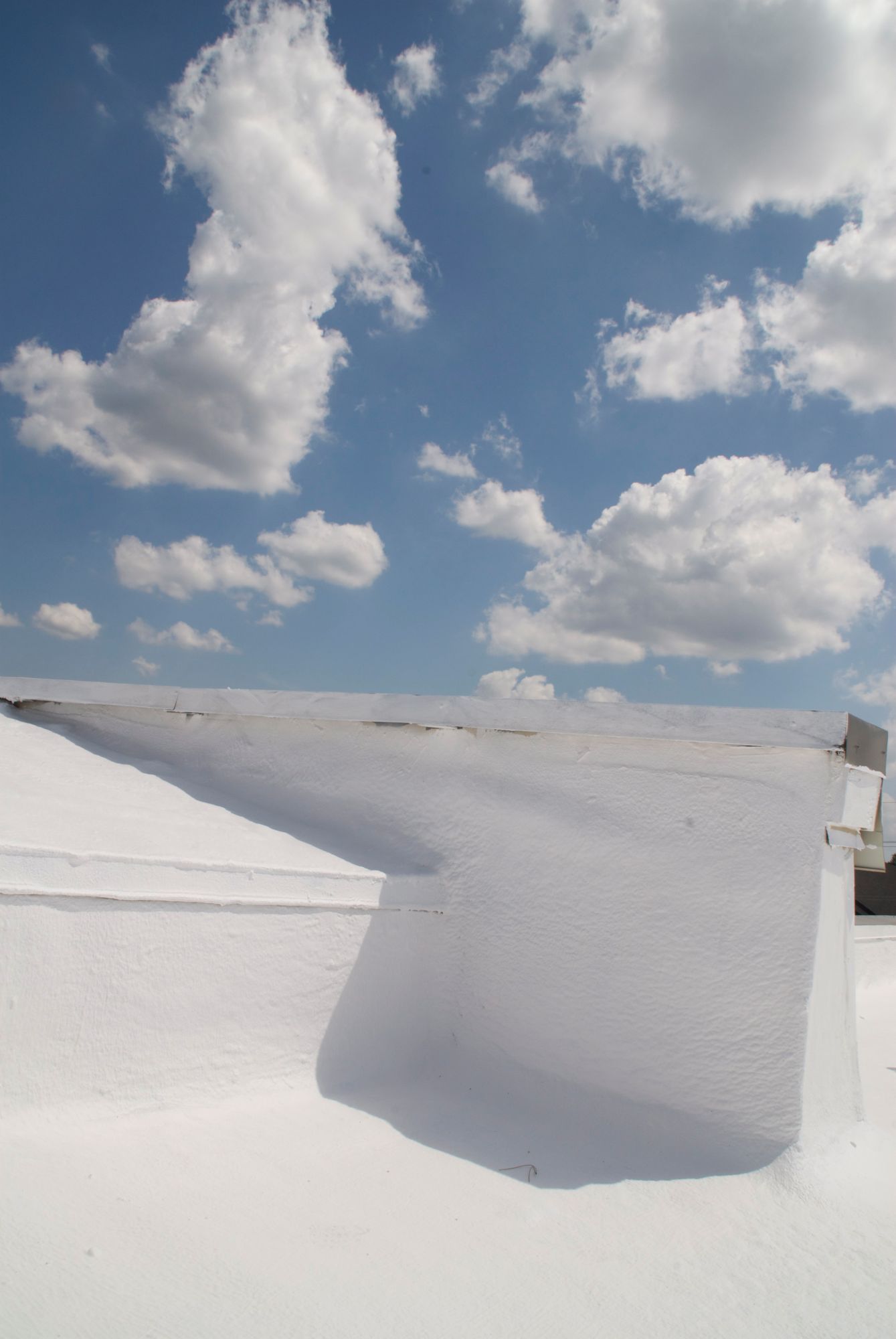 A white, textured architectural structure casts a sharp shadow under a vibrant blue sky filled with scattered white clouds.