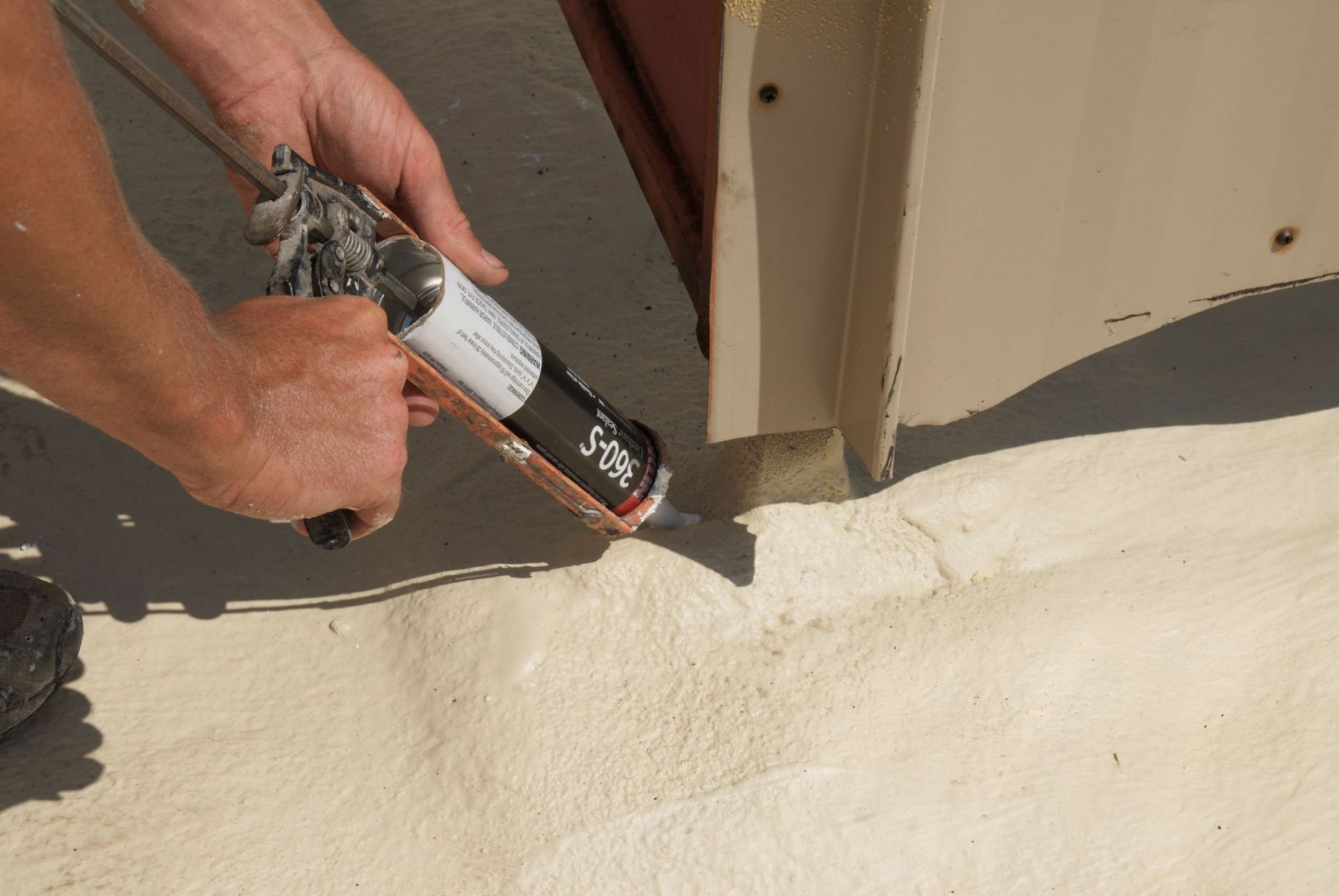 Hands using a caulk gun to apply sealant along the base of a metal structure on a light-colored roof.