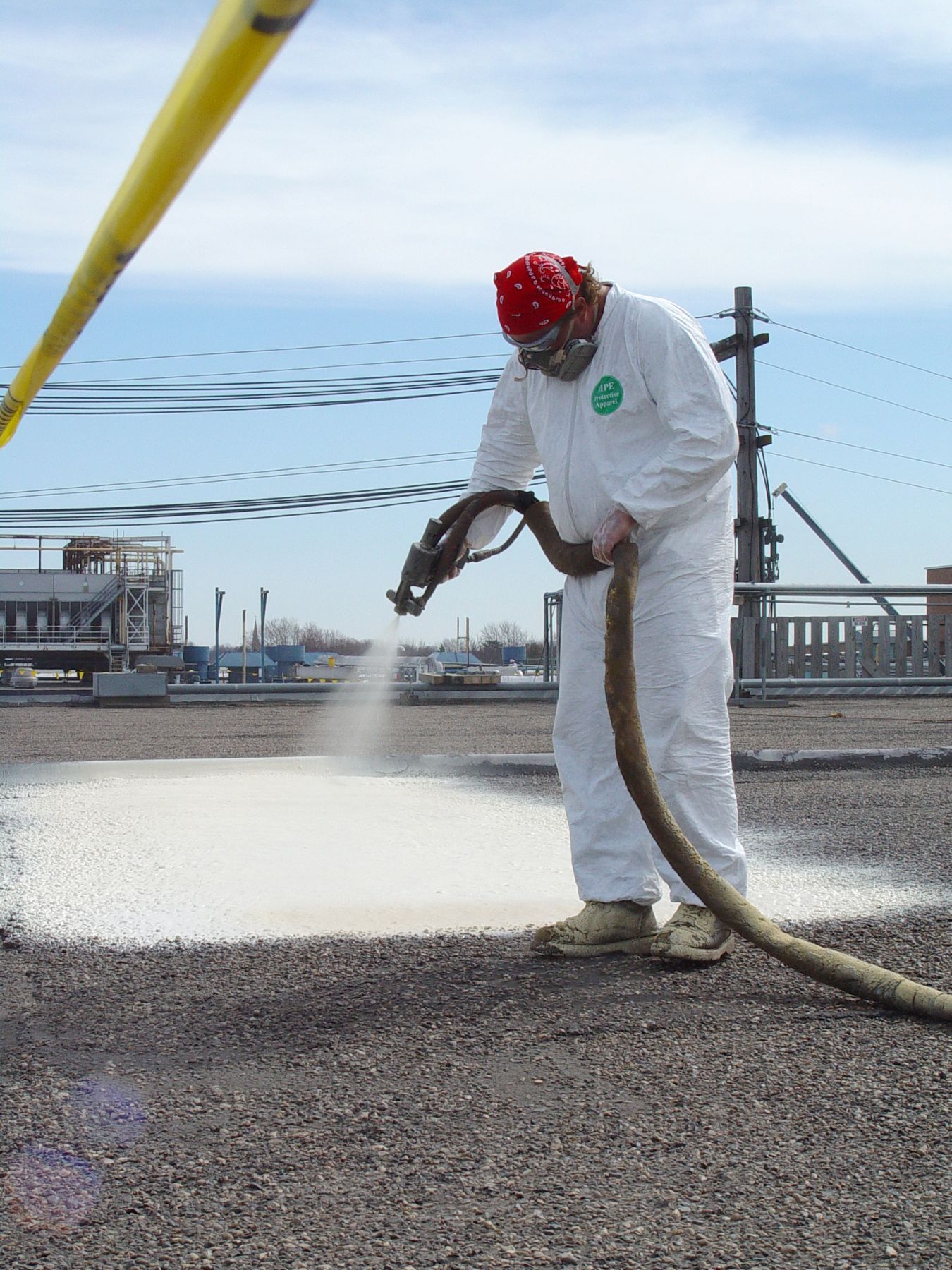 A worker in a white protective suit and red bandana sprays white coating onto a flat roof outdoors.