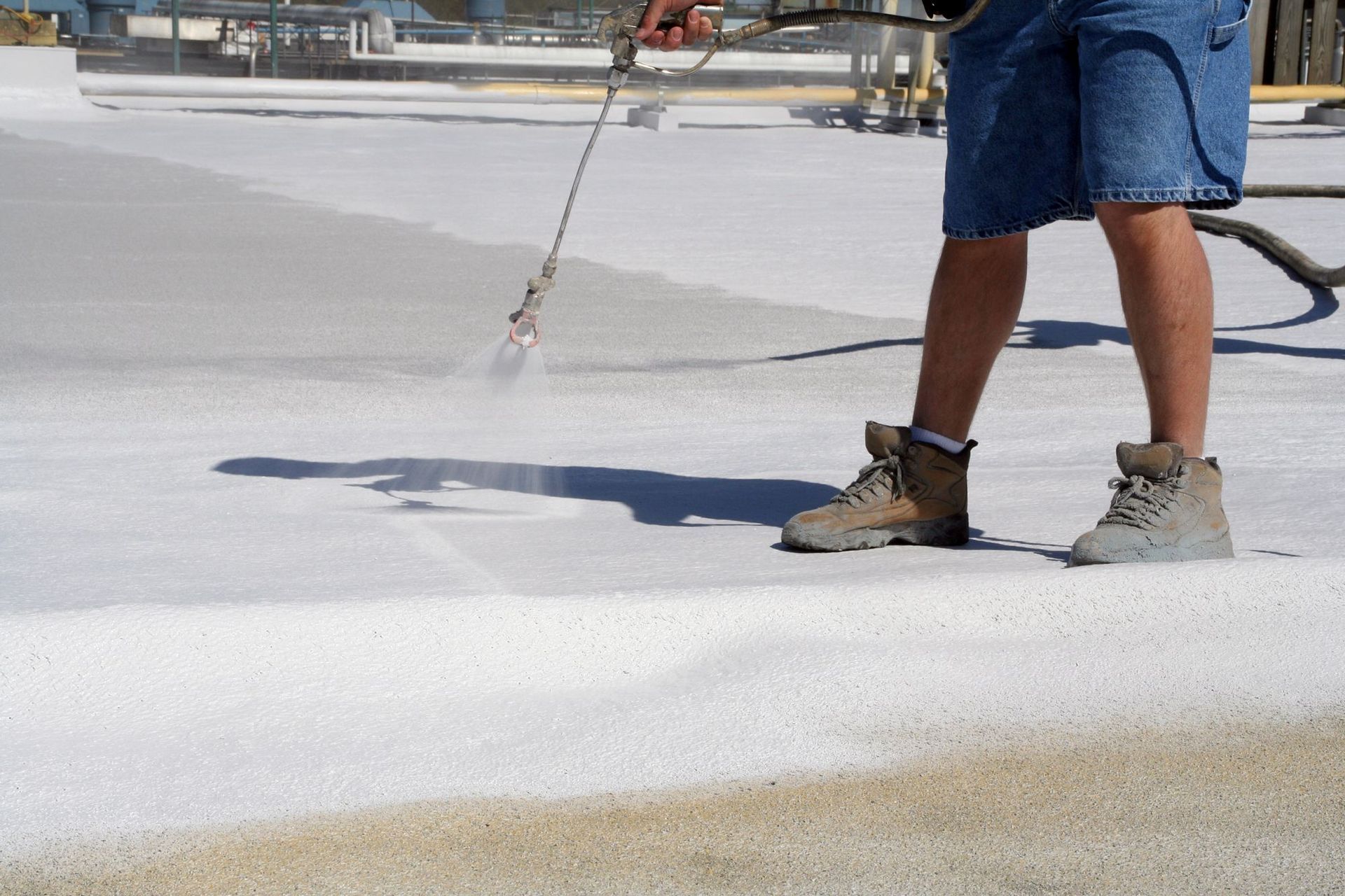 A person in work boots sprays a white, reflective coating onto a flat roof surface.