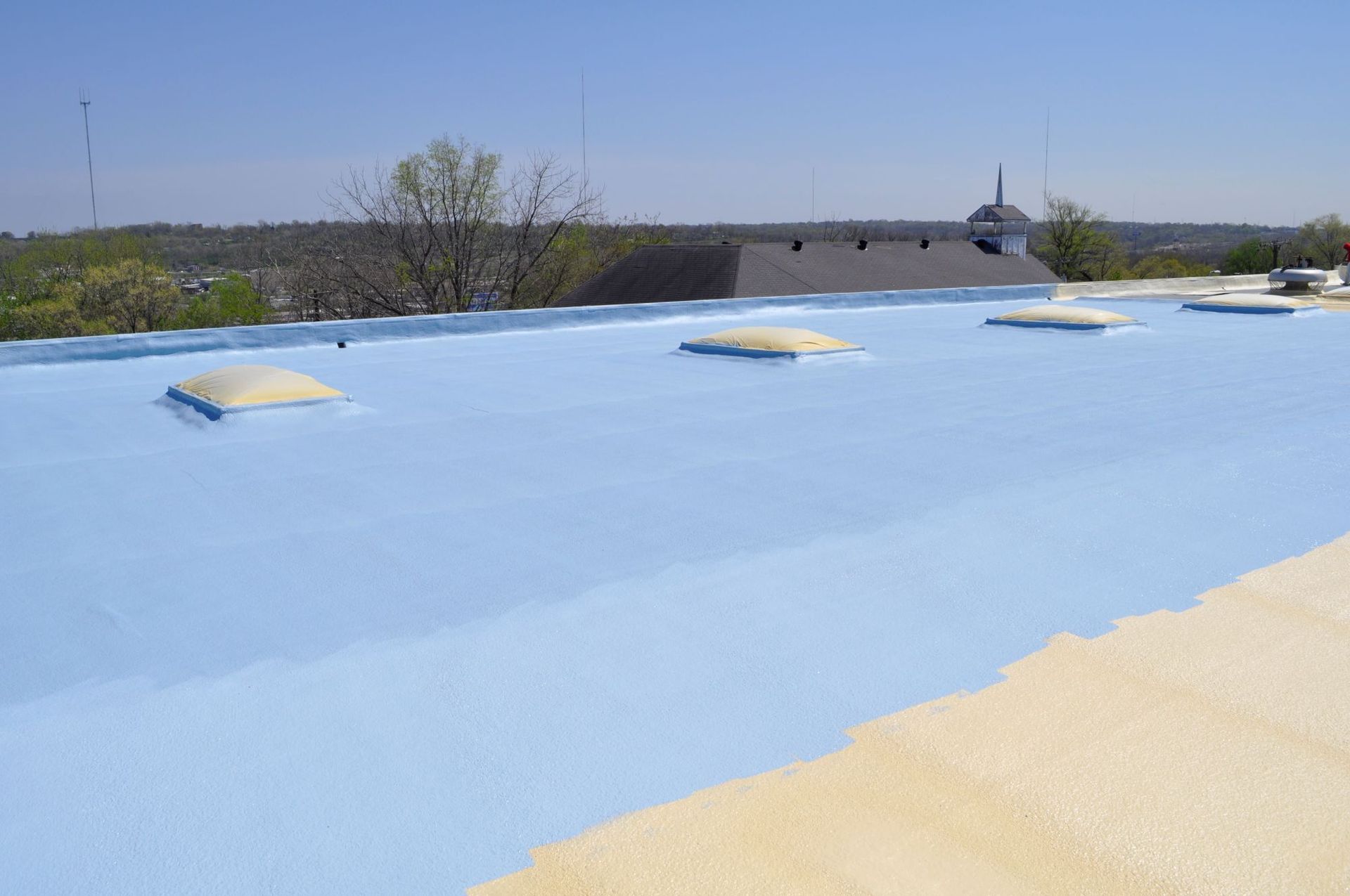 A flat roof partially coated in blue sealant, with several skylights against a clear blue sky.