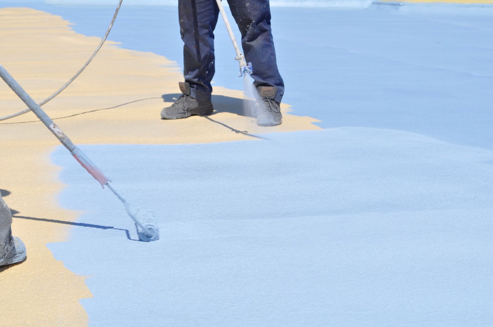 A person uses a roller to apply a light blue protective coating onto a flat, tan-colored roof surface.