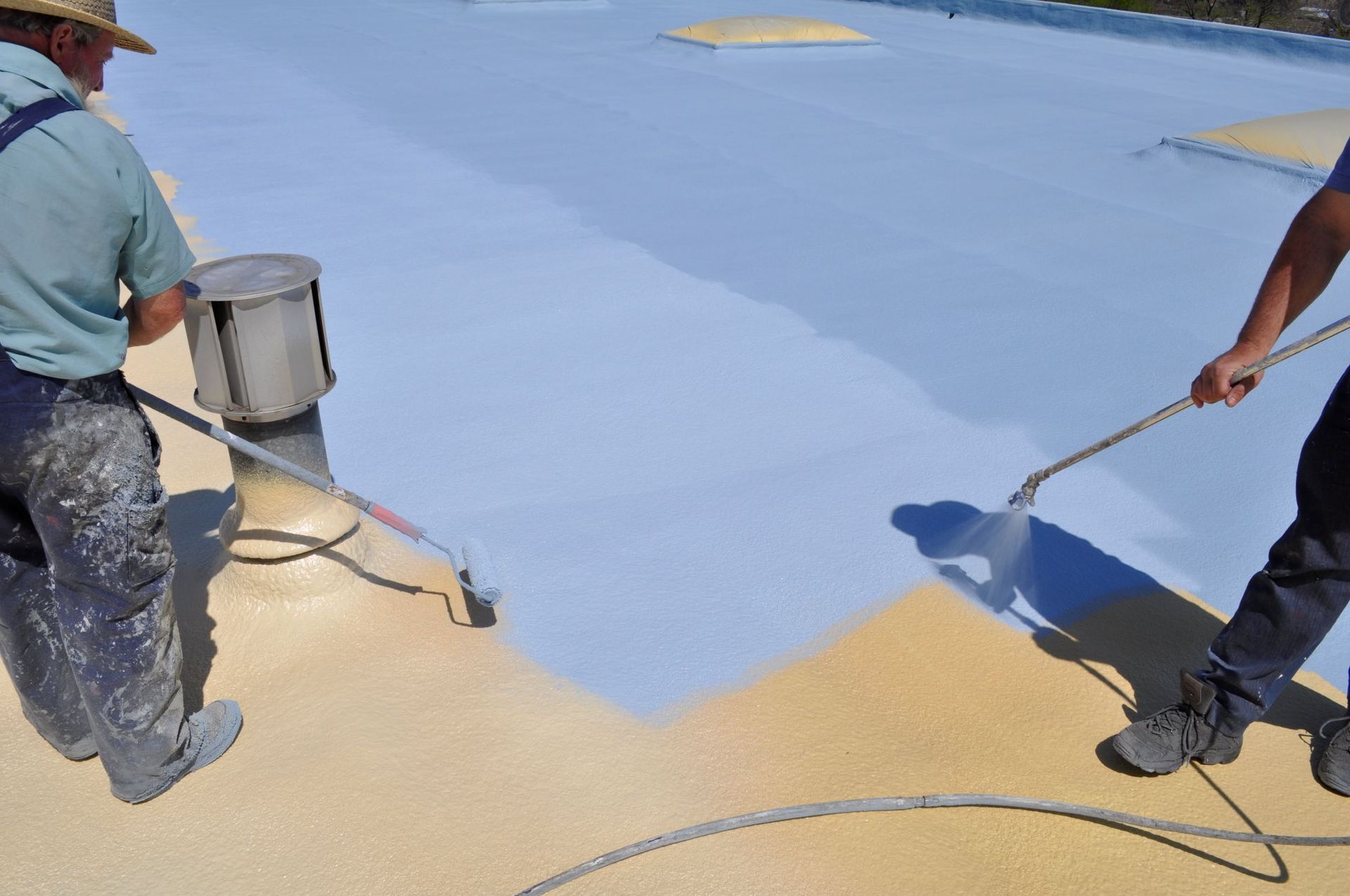 Two workers use sprayers to apply a light blue protective coating to a flat roof, covering the existing yellow surface.