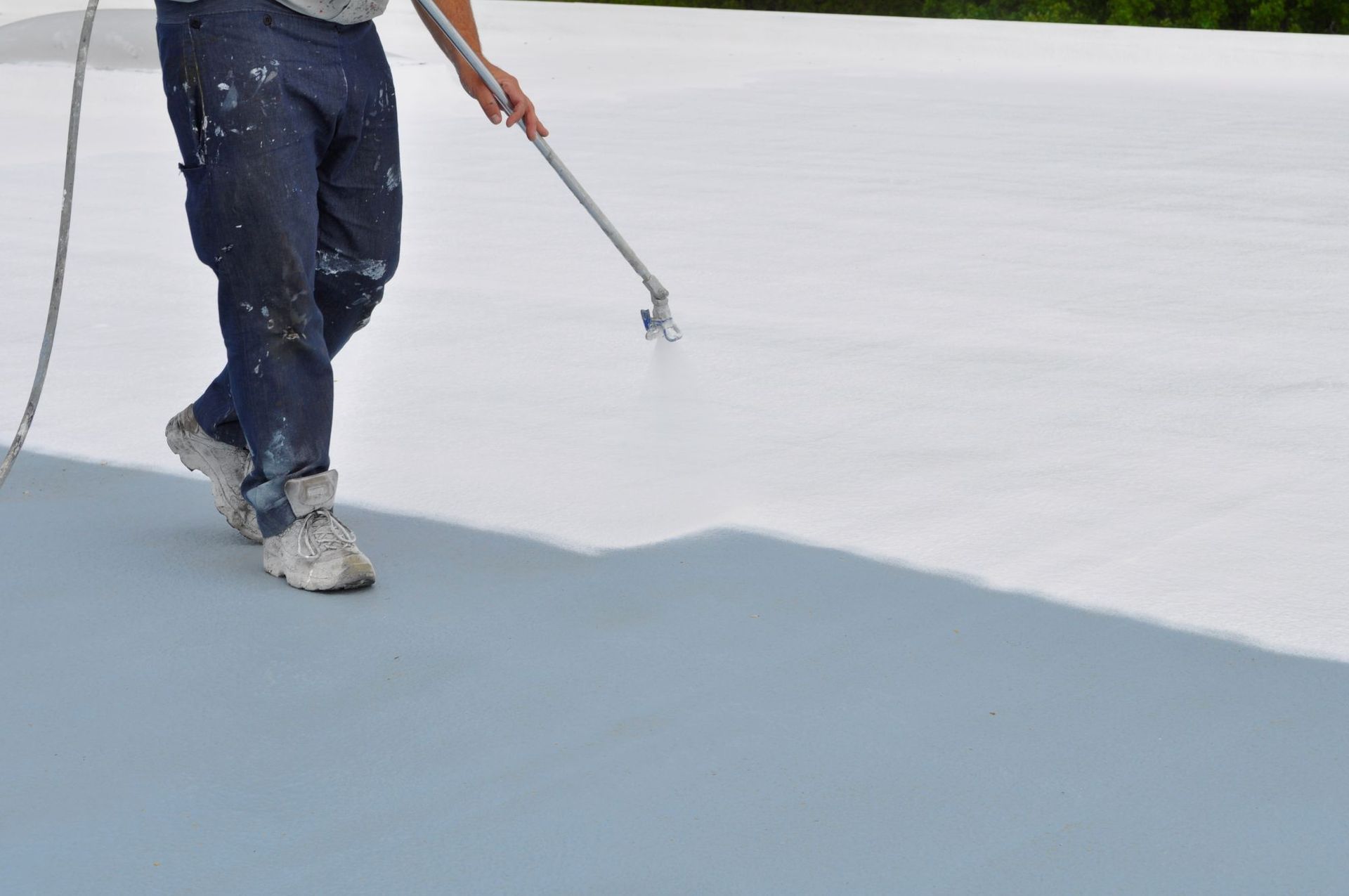 A person using a spray gun to apply a white coating to a gray flat roof.
