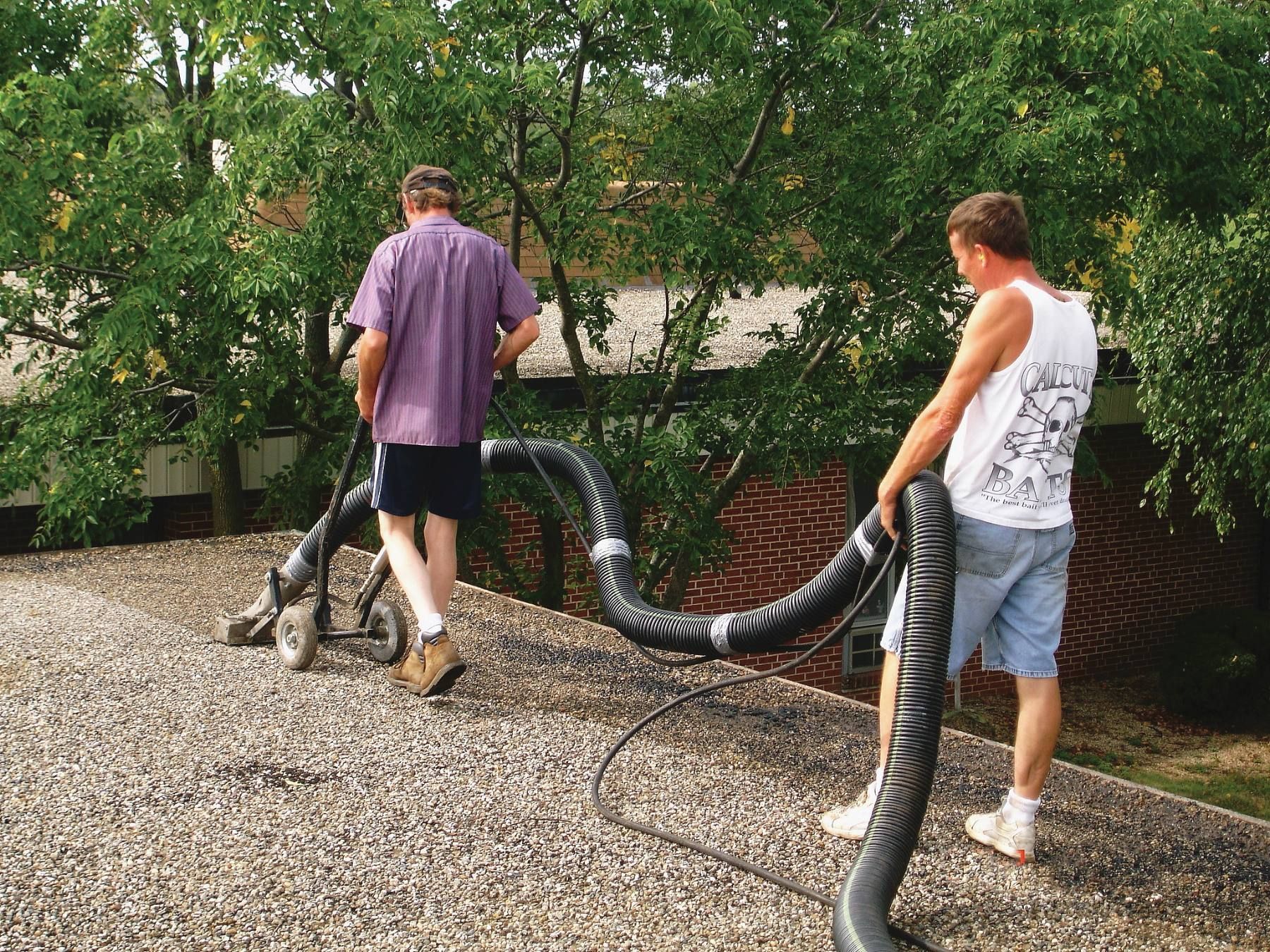 Two workers operate a vacuum hose on a gravel-covered rooftop near trees and a brick building.