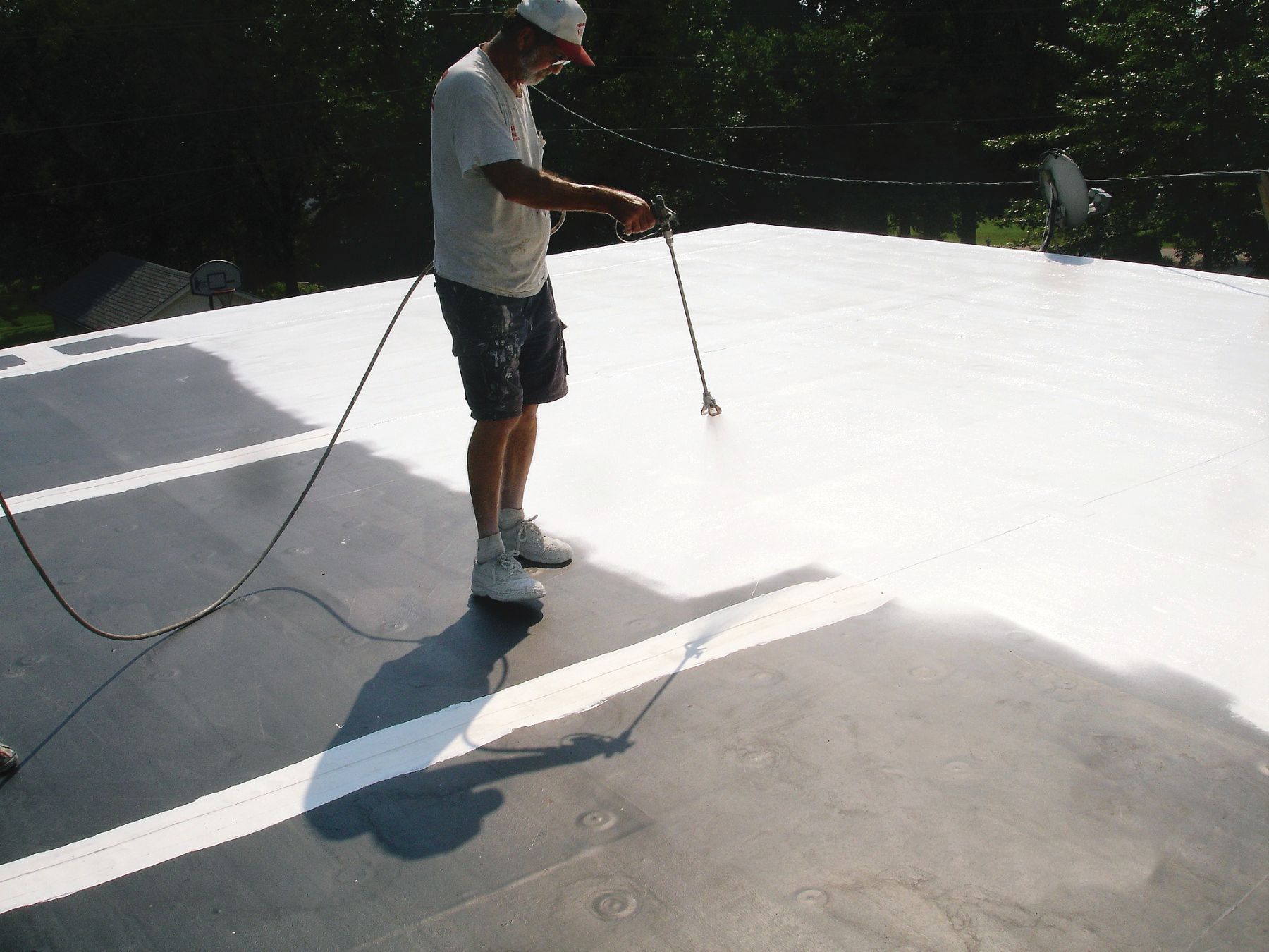 A person uses a spray gun to apply a white reflective coating onto a flat, dark roof surface.