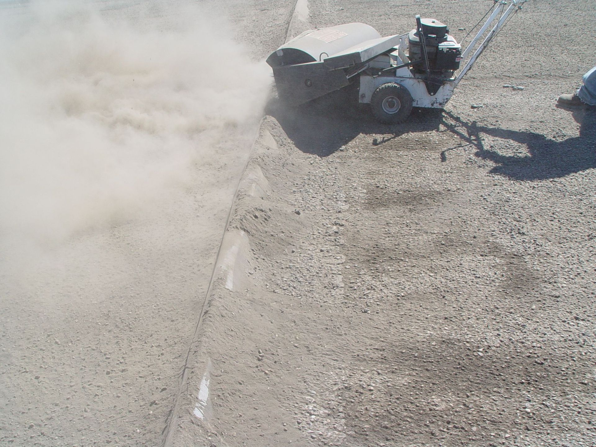 A walk-behind power broom kicks up a cloud of dust while sweeping a gravel-covered outdoor surface.