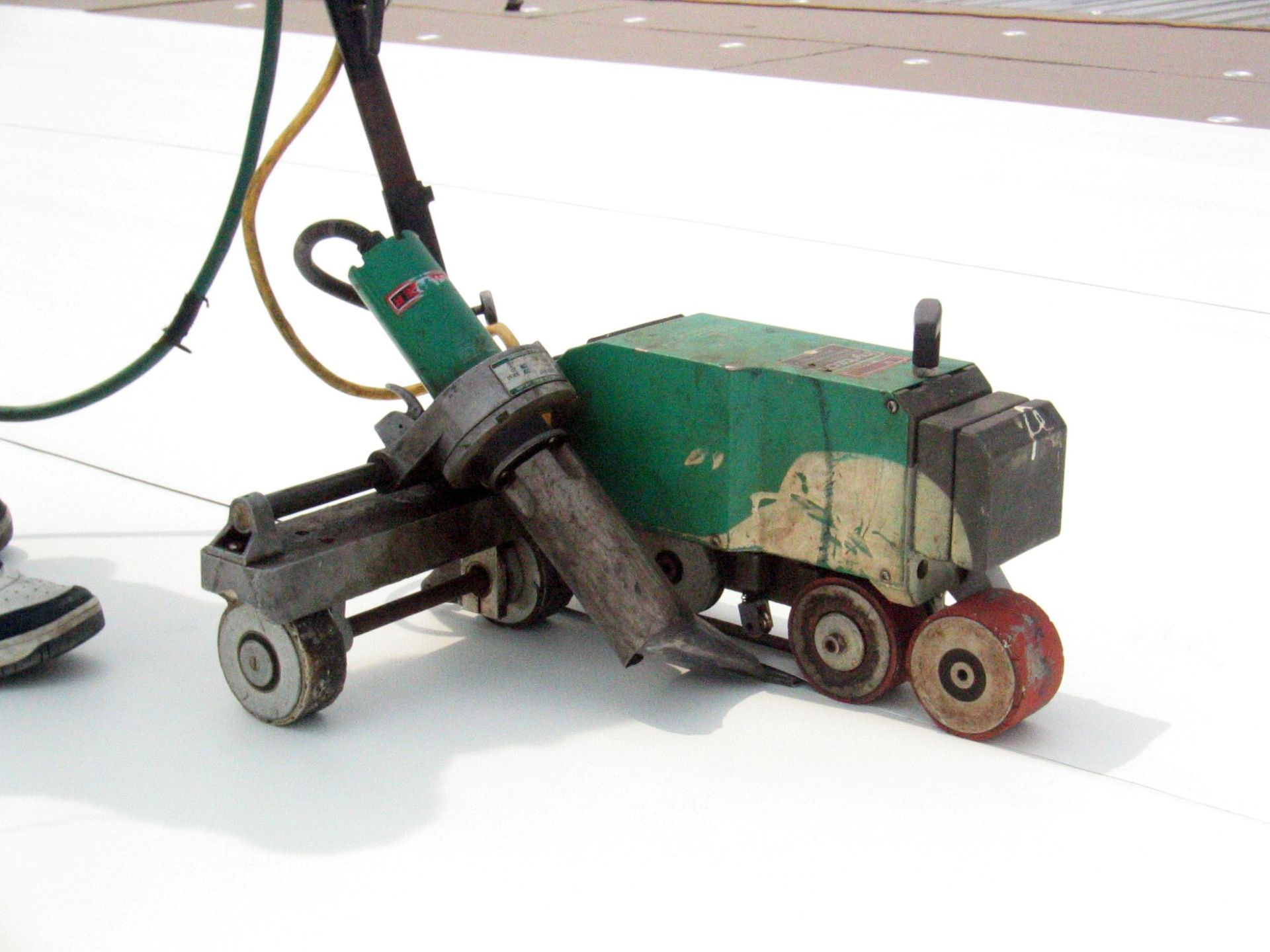 A green automatic hot-air welder machine fuses seams on a white industrial roof membrane.