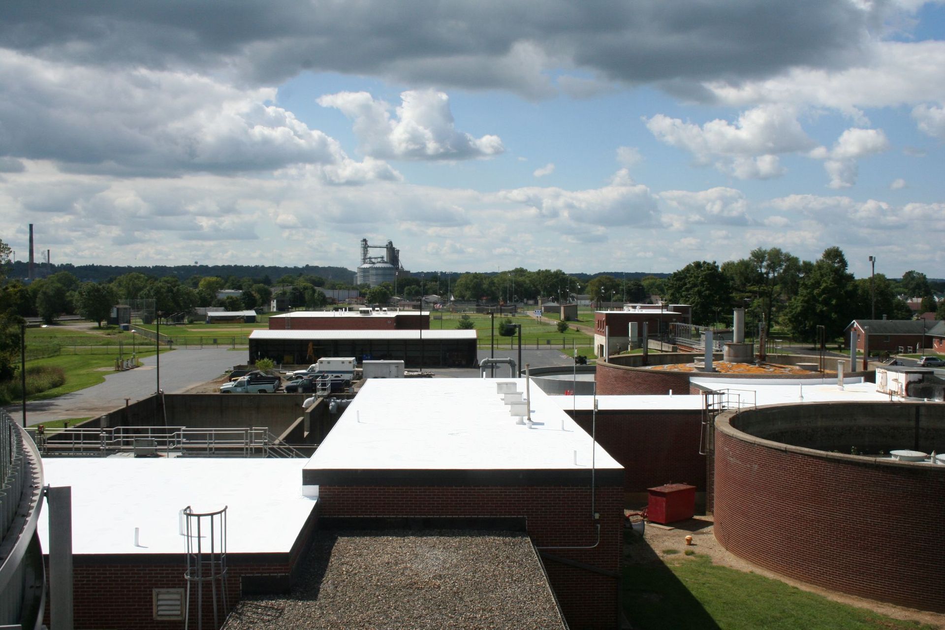 A wastewater treatment facility with circular brick tanks and flat-roofed buildings under a cloudy sky.