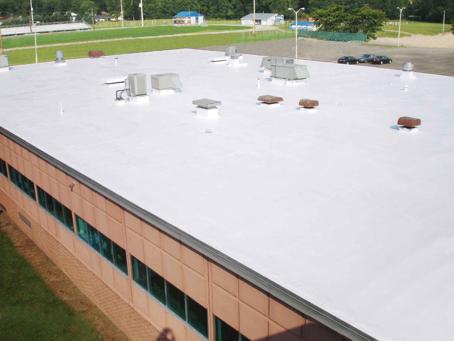 An aerial view of a flat, white commercial roof with HVAC units, bordering a brick building and grassy lot.