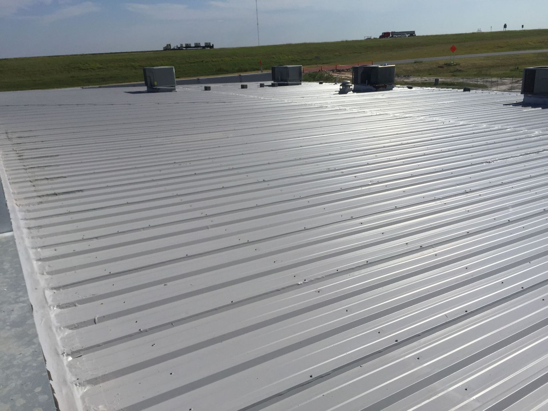 A low-angle view of a flat, light-gray metal roof with several HVAC units against a clear sky and grassy horizon.