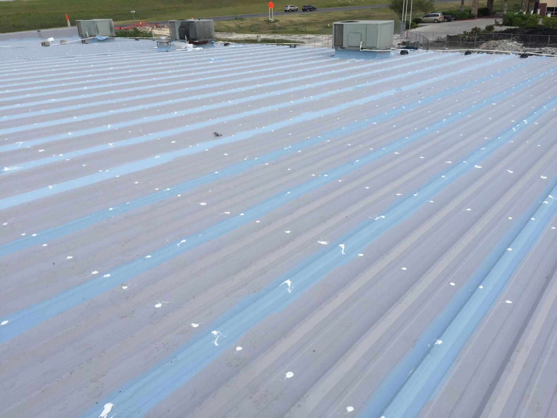 A metal industrial roof with grey panels, blue seam sealants, and white fasteners, seen from a high angle.