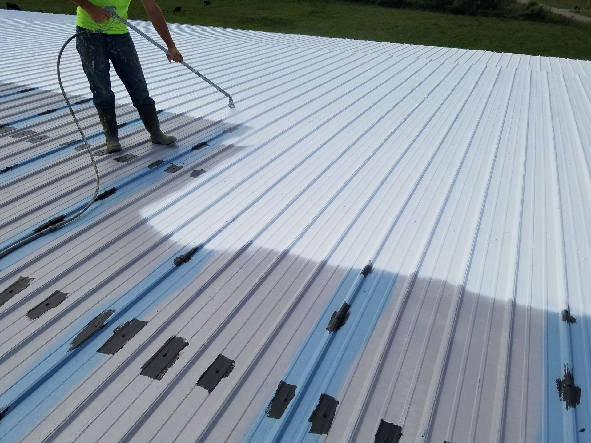 A worker uses a spray gun to apply white reflective coating to a corrugated metal roof.