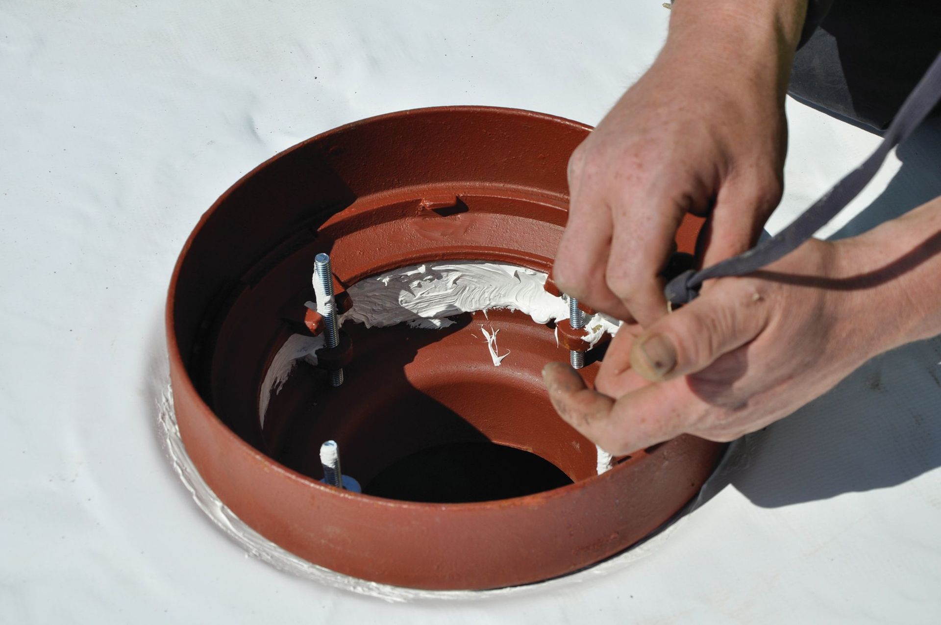 Hands work on a red-brown circular vent fitting being installed on a white flat roof.