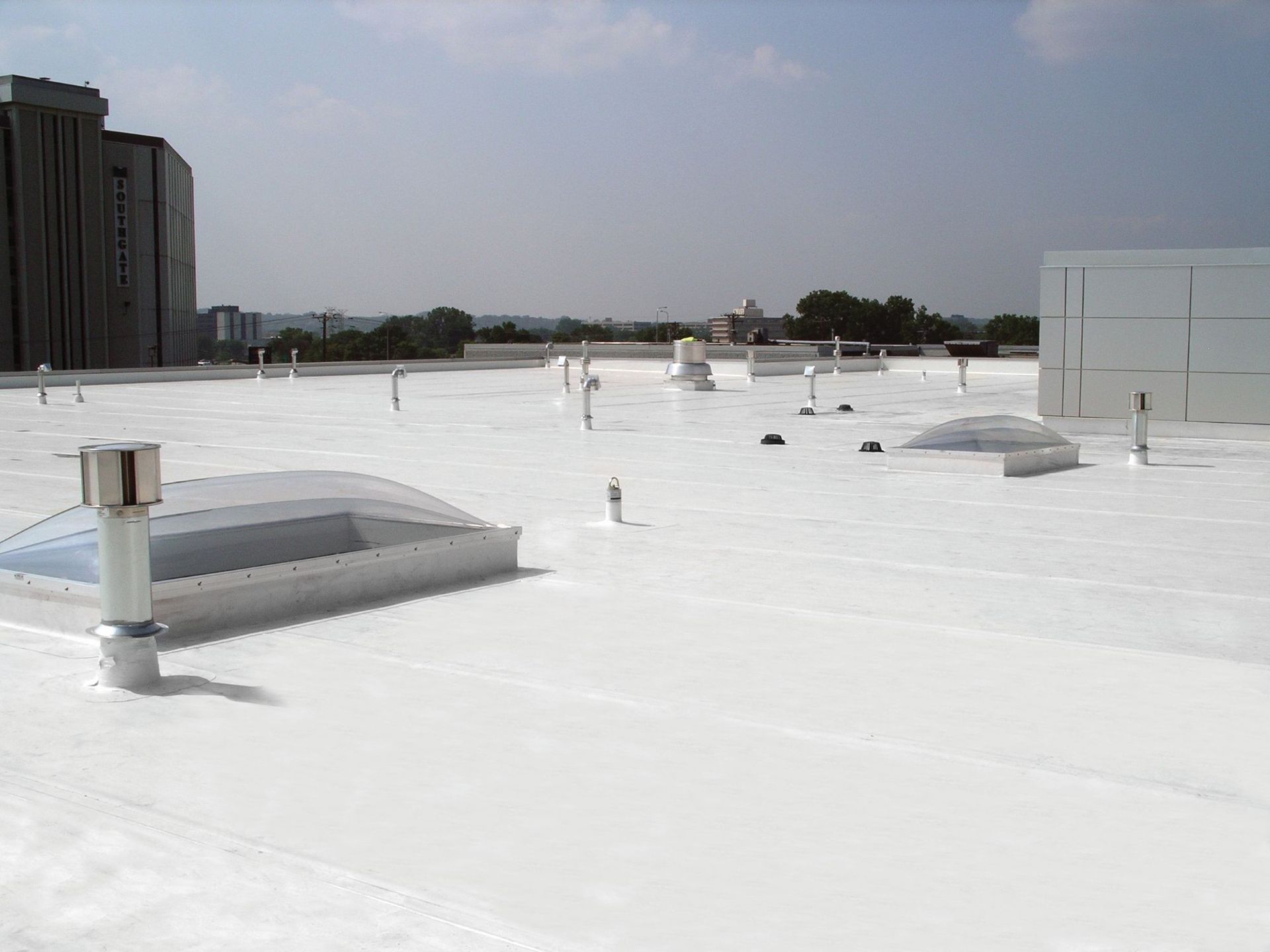 White flat roof with several skylights, ventilation pipes, and a building in the background under a clear sky.