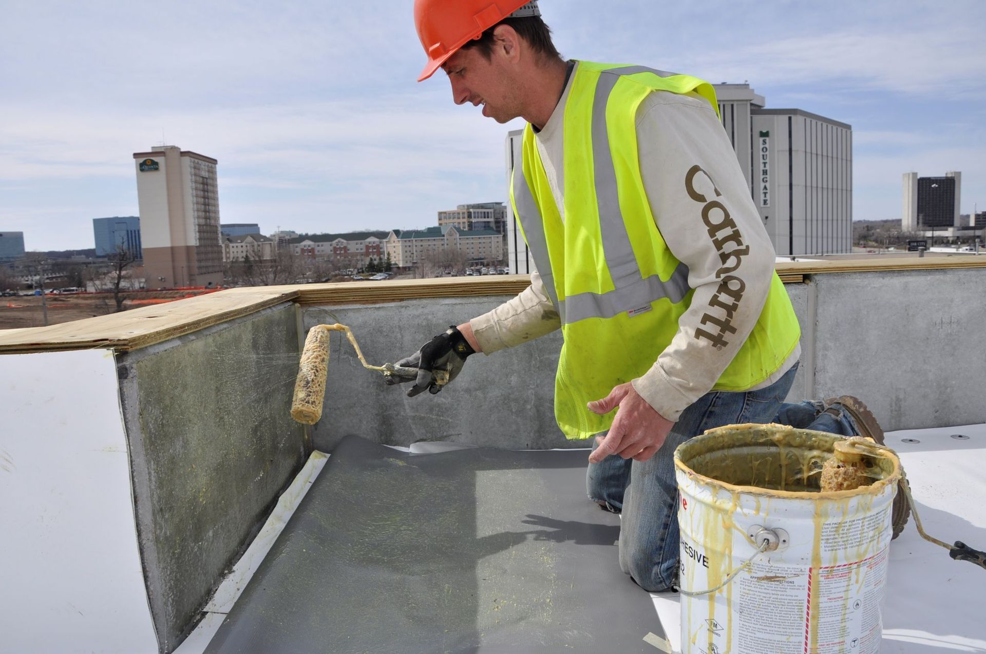 A construction worker in a hard hat and safety vest applies adhesive to a rooftop wall with a paint roller.