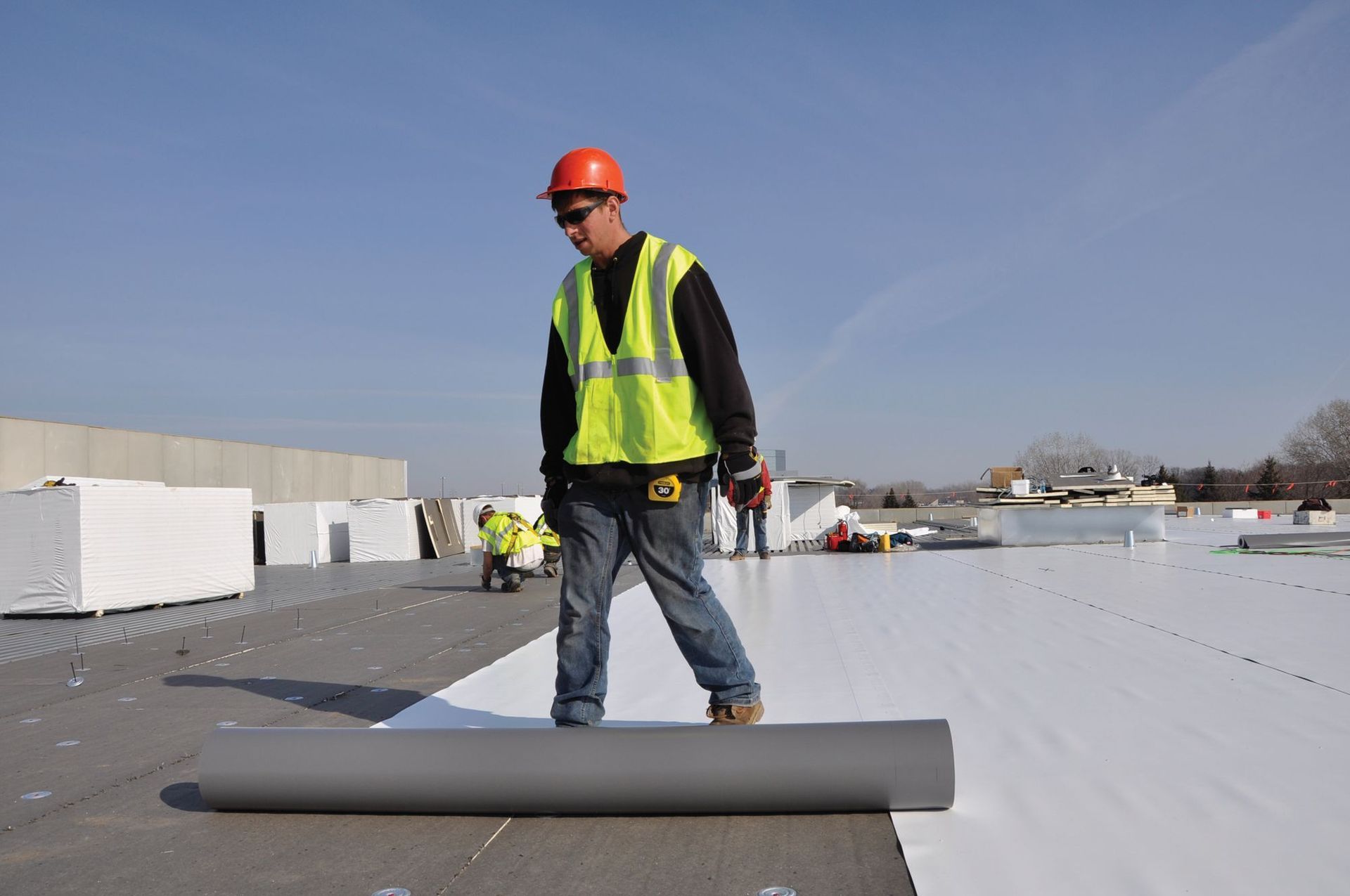 A worker in a high-visibility vest and hard hat unrolls grey roofing material on a sunny rooftop construction site.