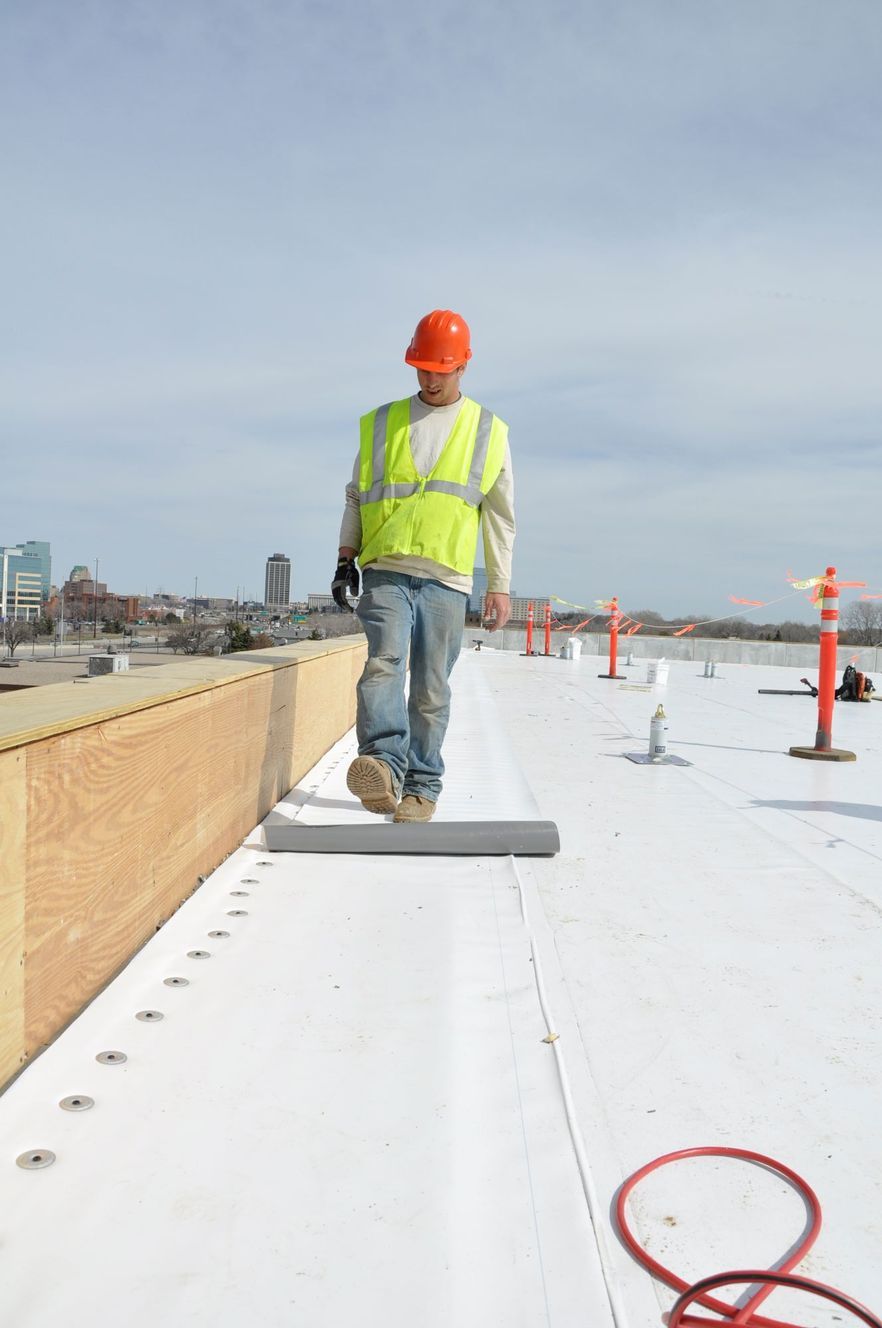 A worker in a high-visibility vest and hard hat installing roofing material on a flat roof with an urban background.