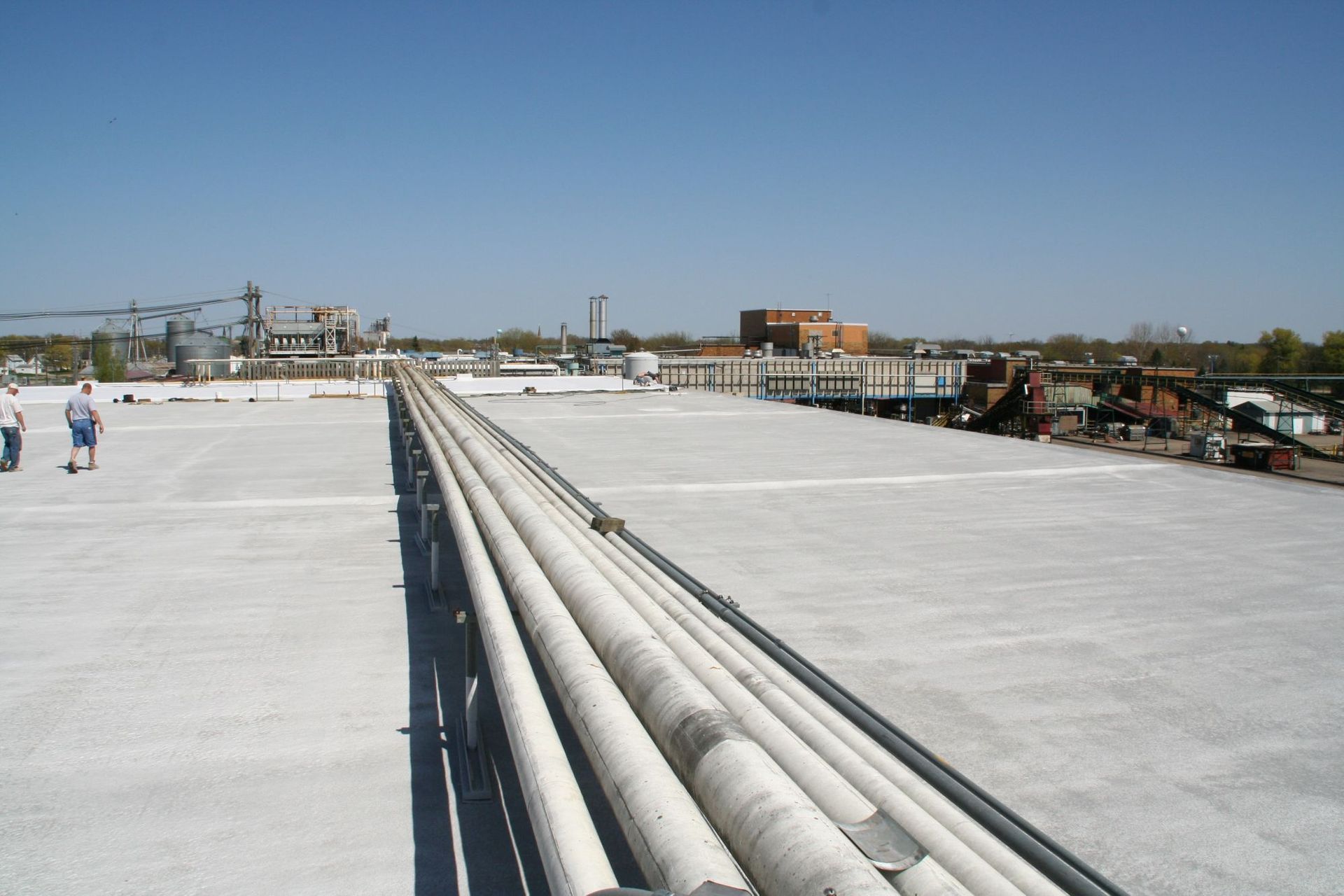 Two people walk on a wide, light-gray industrial rooftop next to a series of long, parallel pipes under a clear blue sky.