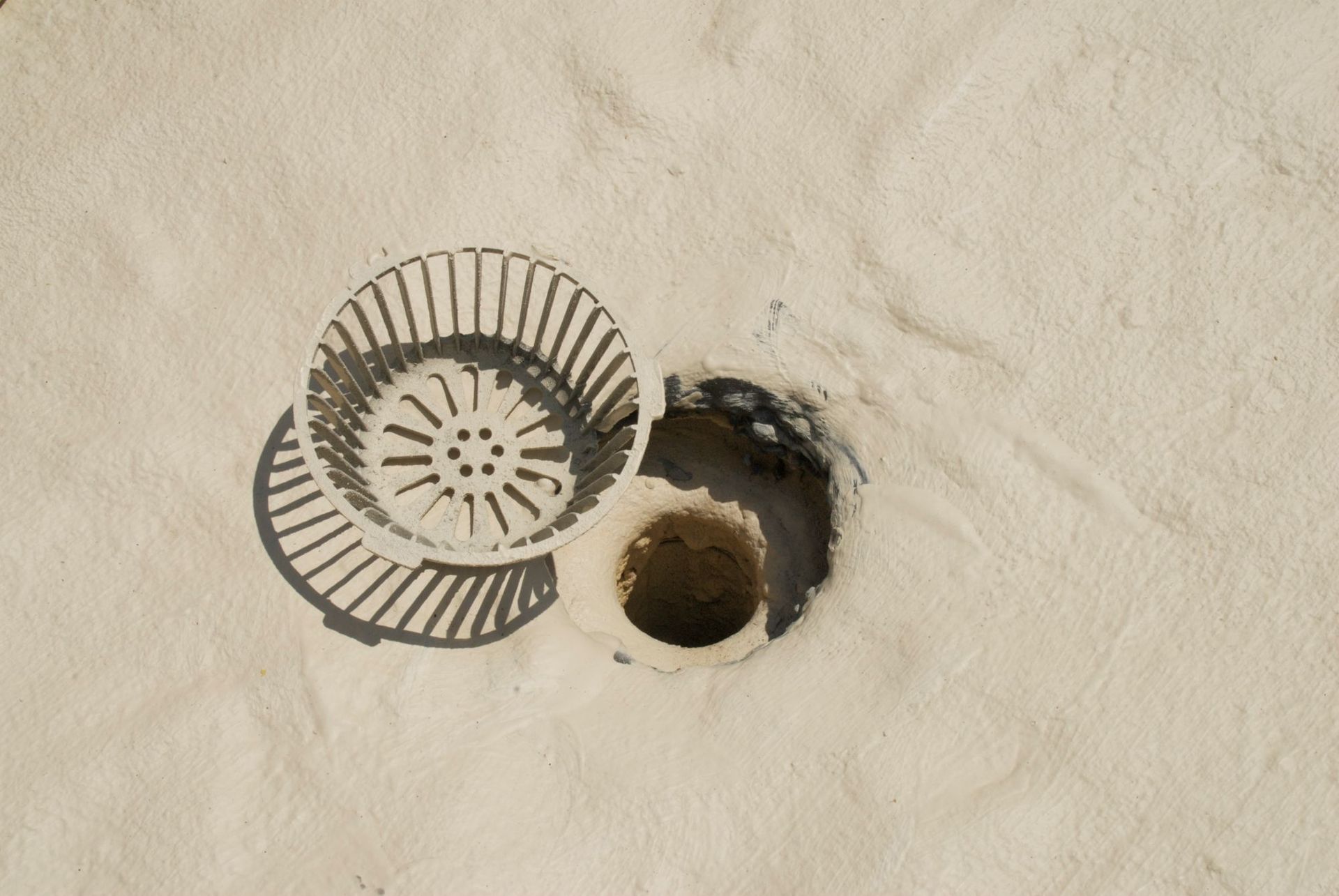 A plastic drain cover sits detached next to an open, circular hole in a light-colored, textured concrete surface.