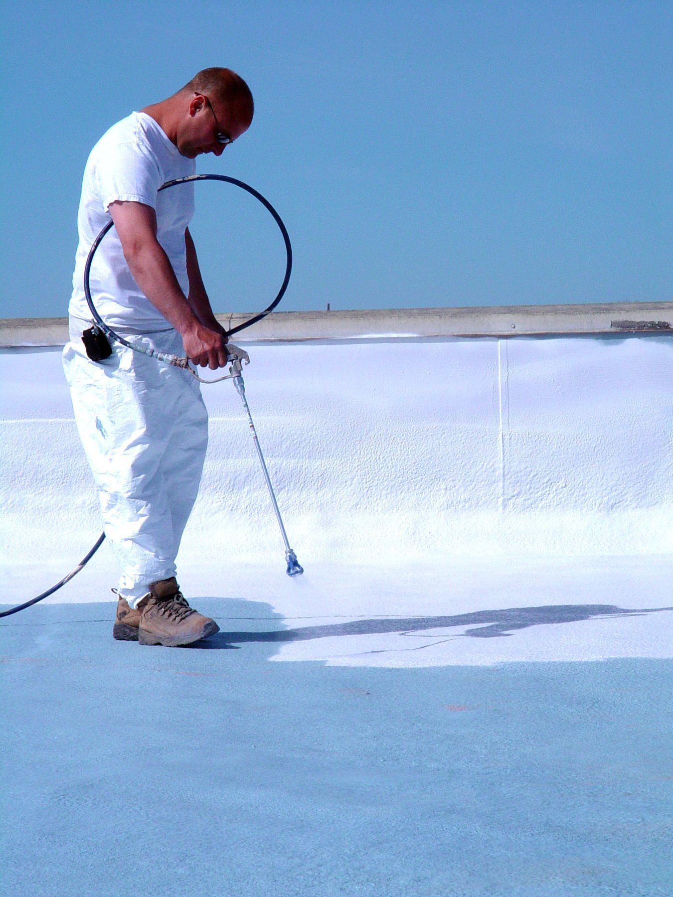 A worker in white protective gear sprays a white sealant onto a flat roof on a clear, sunny day.