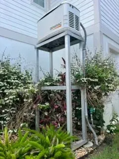Outdoor HVAC unit raised on a tall metal platform beside a house and dense garden foliage