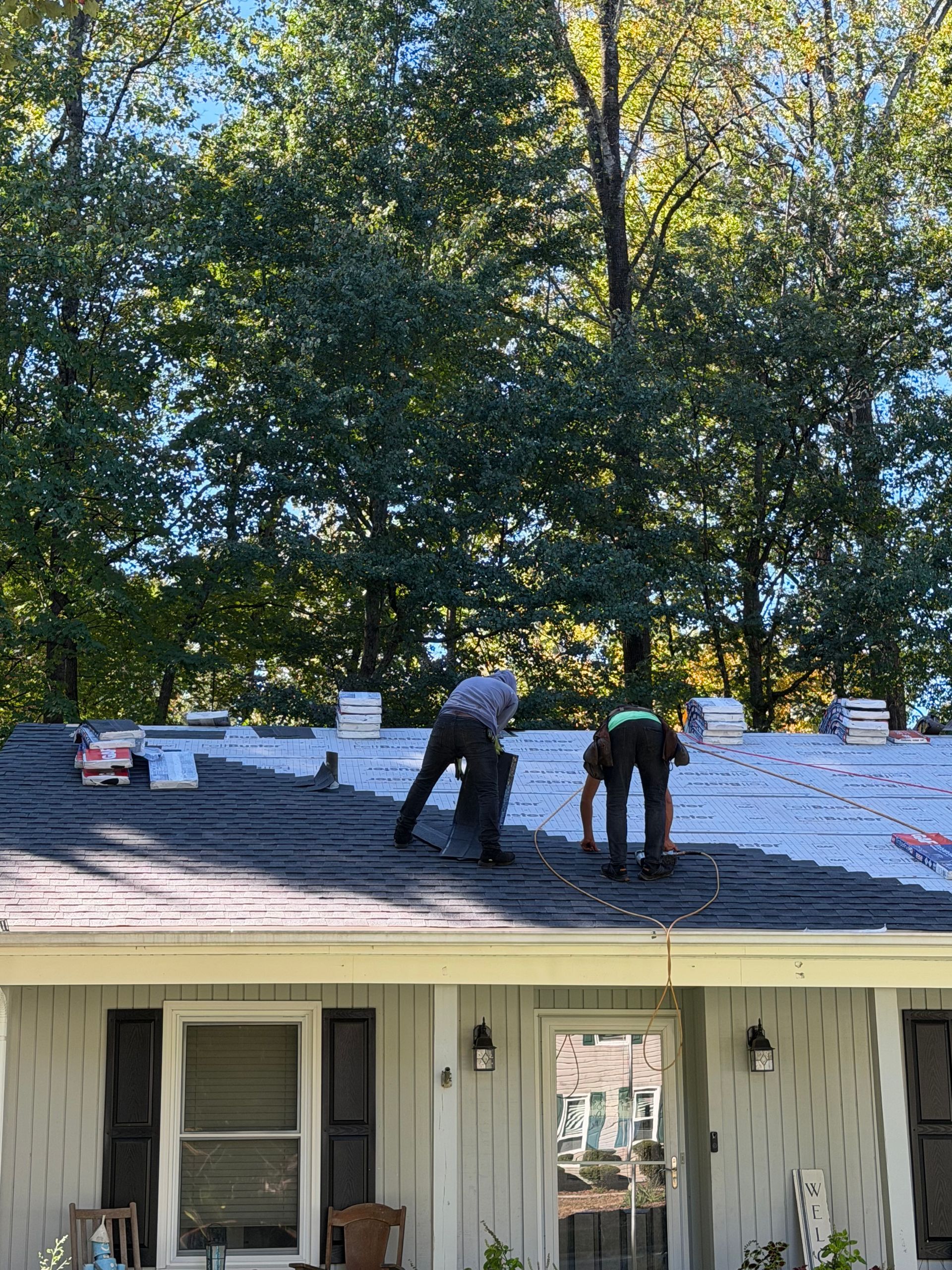 Two workers install black shingles on a house roof on a sunny day with trees in the background.
