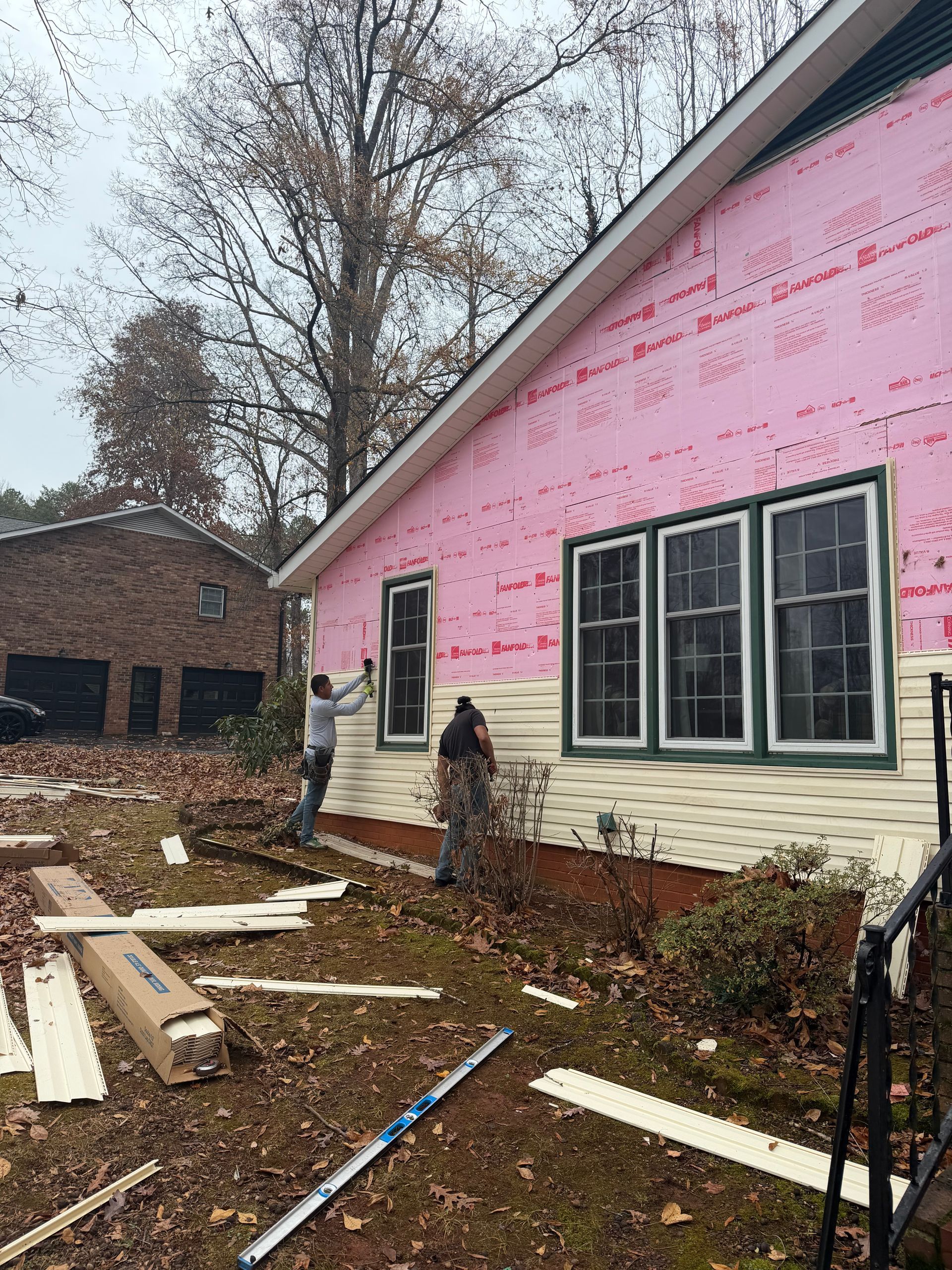 Two workers install light-colored siding on the side of a house with pink insulation visible.