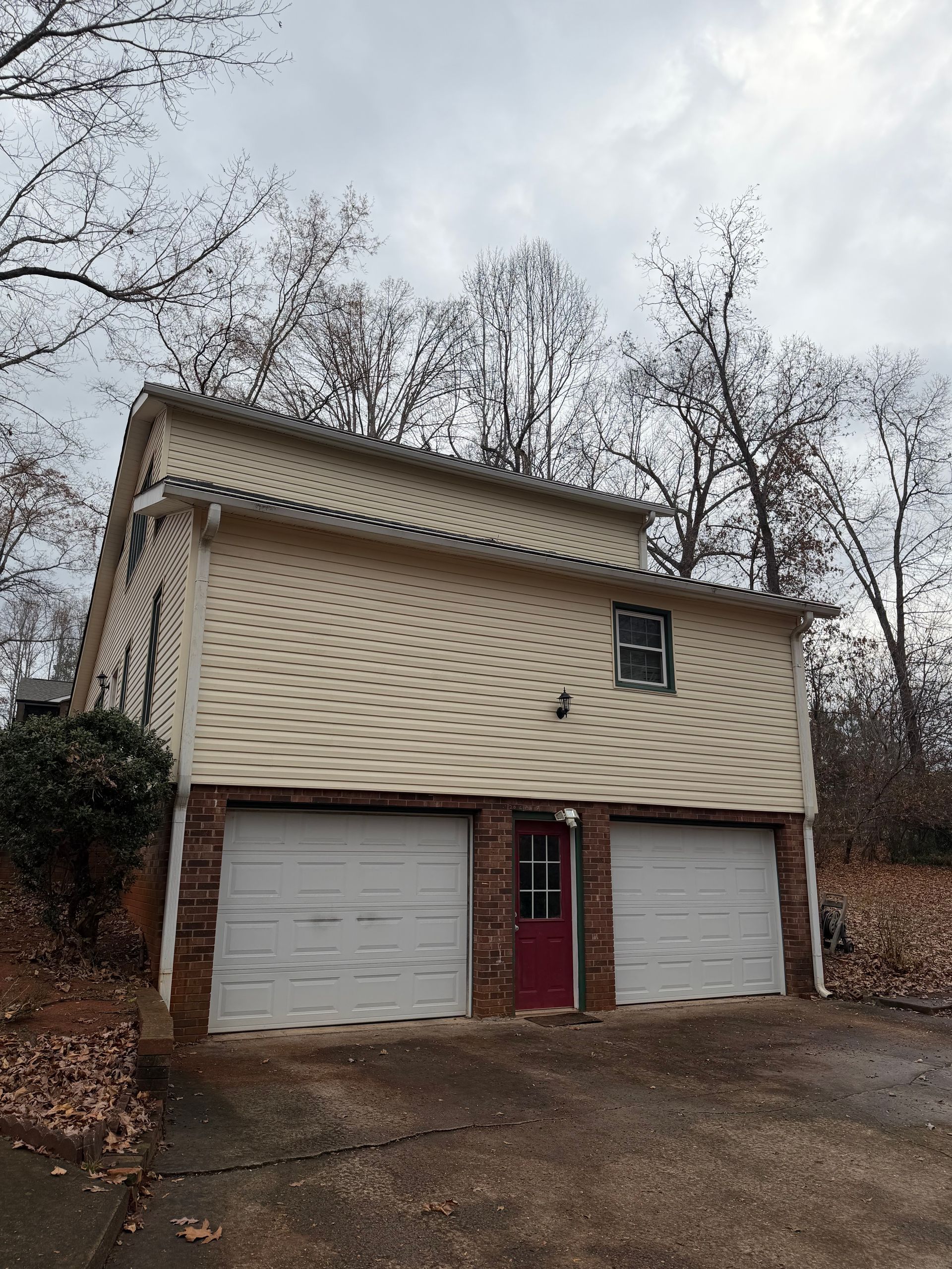 A two-story building with beige siding featuring two garage doors and a red door, set against a backdrop of trees.